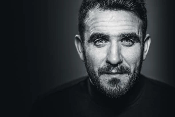 Portrait de studio en noir et blanc d'un homme aux cheveux et à la barbe courts, portant une chemise sombre, regard direct, arrière-plan flou.