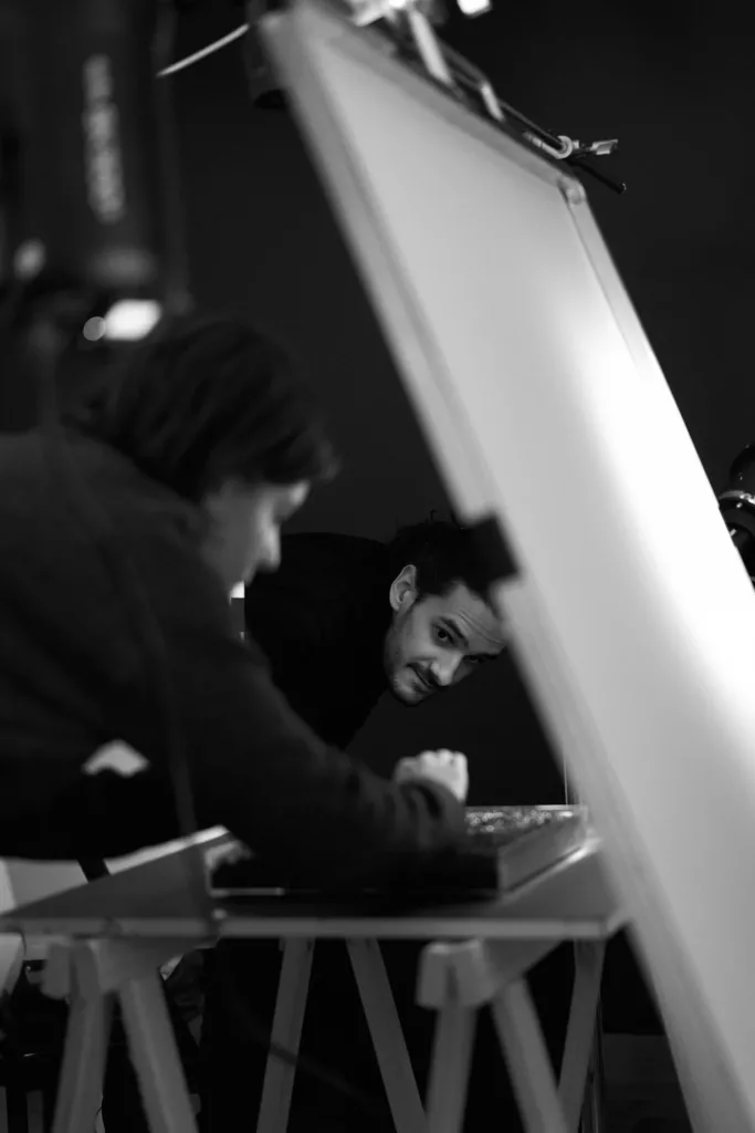 Le photographe qui regarde ajuste son installation à une table de travail en studio ; une deuxième personne fait face à l'appareil photo. Un éclairage de studio et un panneau réflecteur sont présents.
