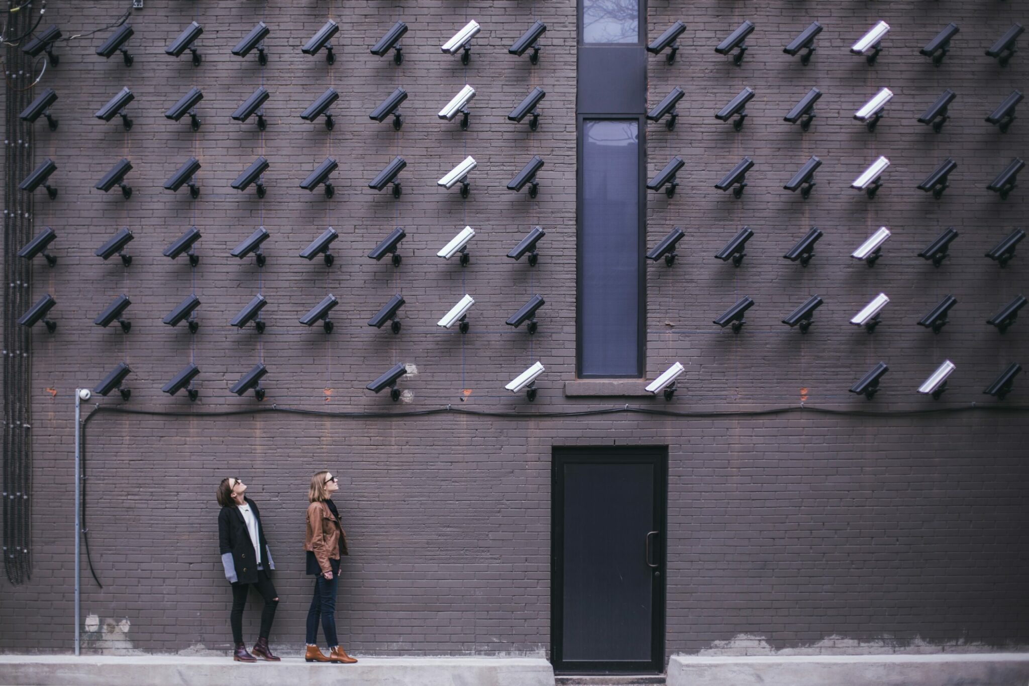 Deux personnes sur un trottoir observent un mur de briques bordé de caméras de sécurité, avec une haute fenêtre et une porte noire ; photographie de rue urbaine.