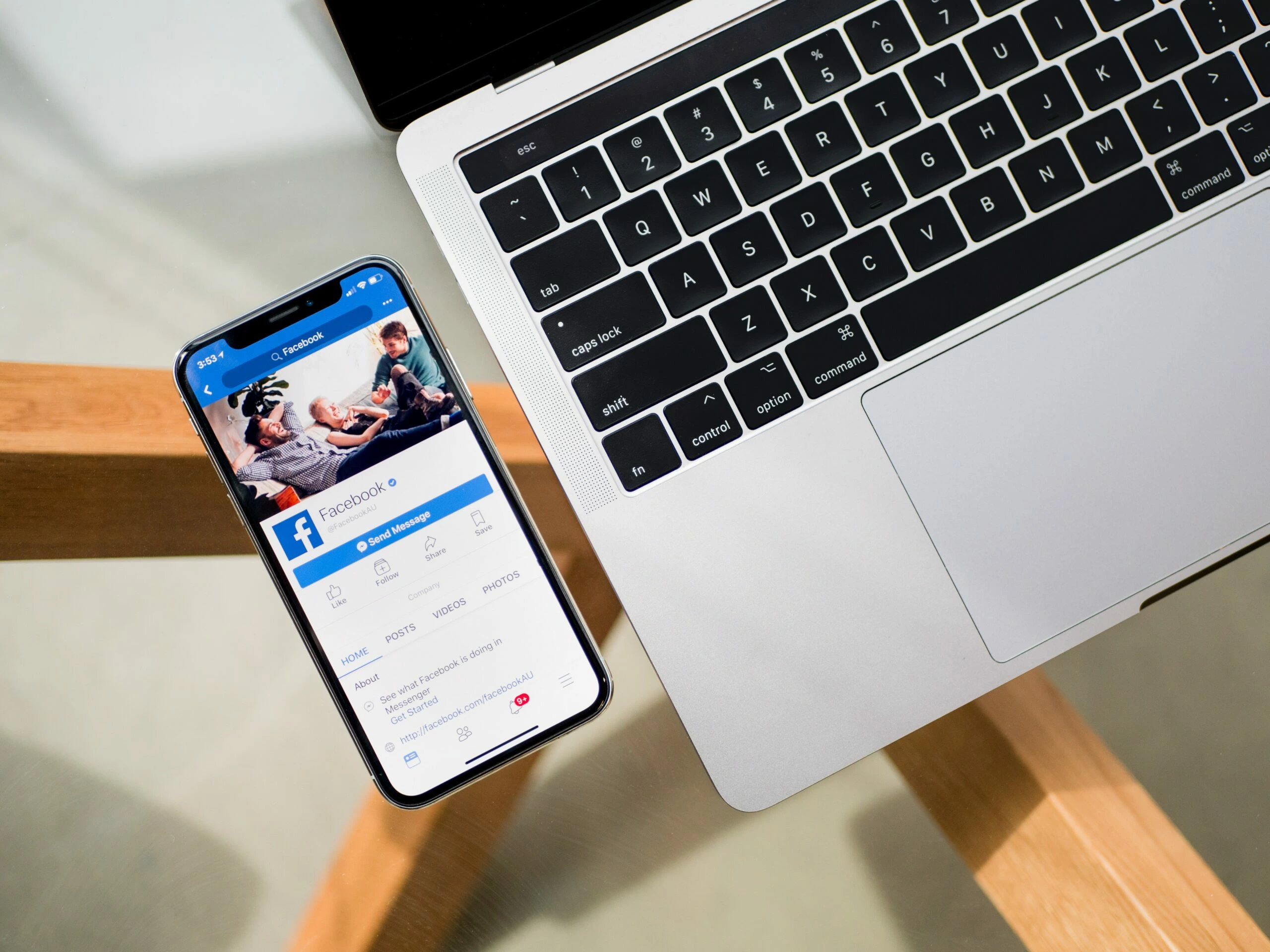 agence de photographie rétines – reseaux-sociaux-entreprise-3 Un smartphone affichant un profil Facebook est posé sur une table en bois à côté d'un clavier d'ordinateur portable ouvert, idéal pour la photographie numérique de produits.