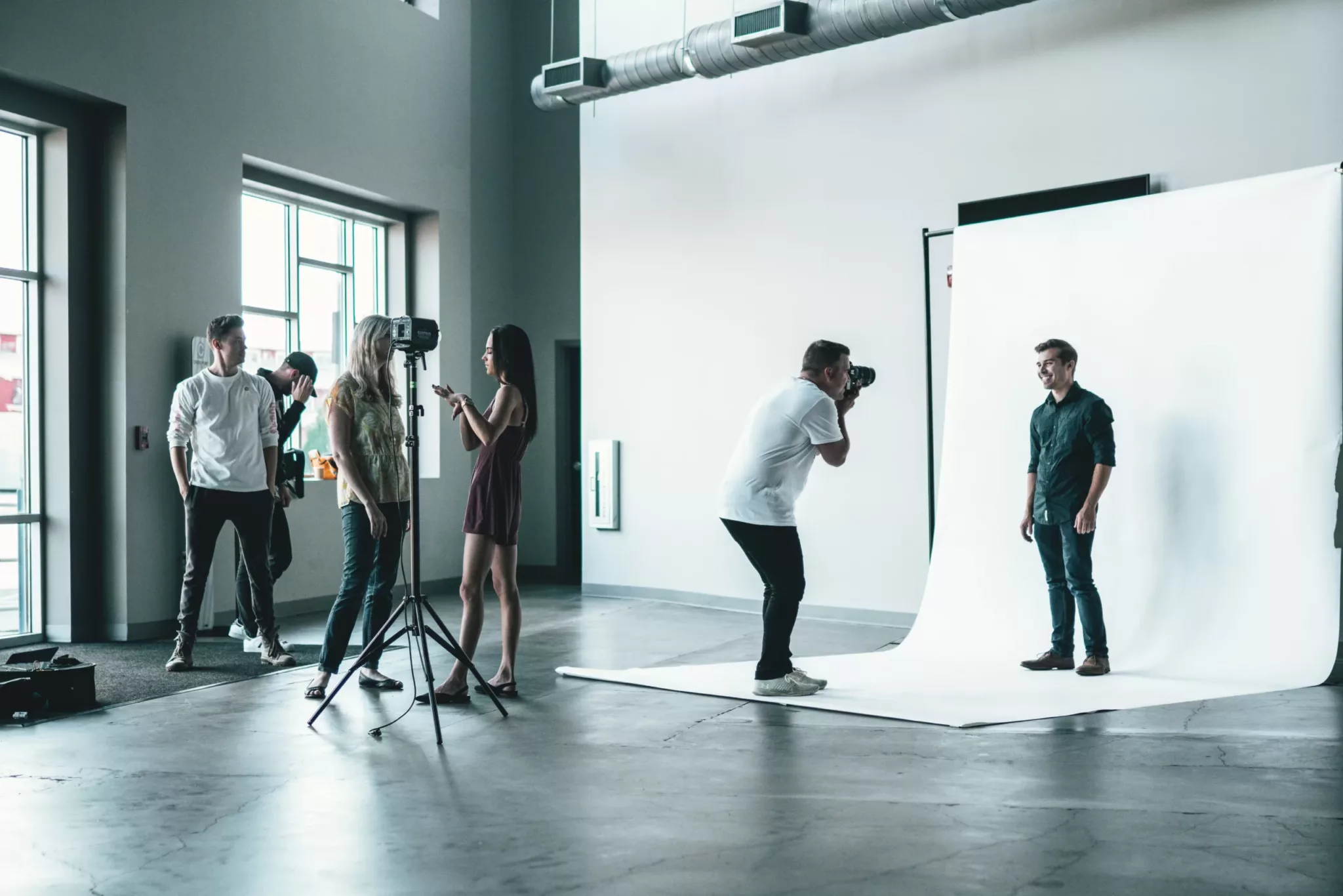 Dans un studio, un photographe prend des photos d'un homme sur un fond blanc, tandis que des assistants règlent l'éclairage et que des employés se concertent près de la fenêtre.