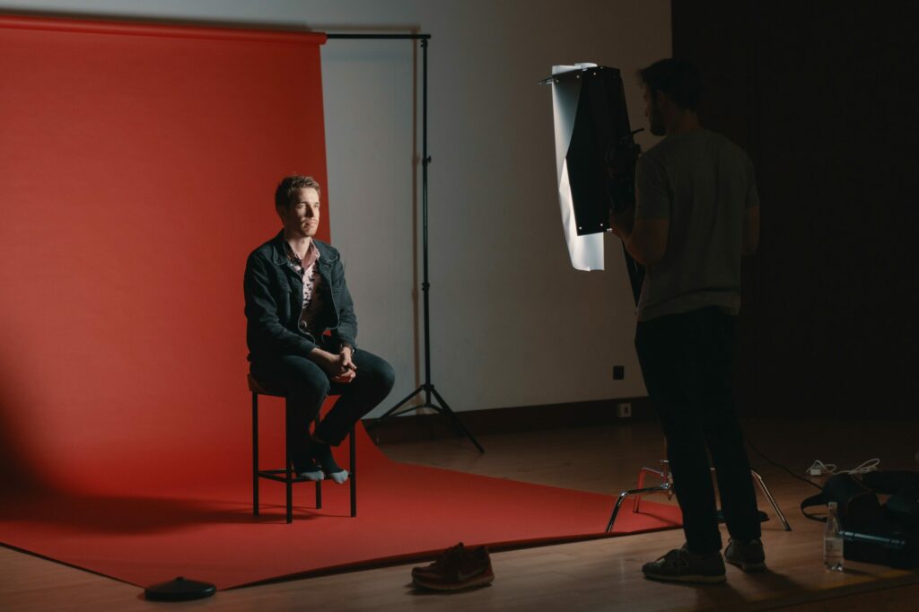 Un homme est assis sur un tabouret face à l'appareil photo sur un fond rouge. L'éclairage de studio et le photographe sont visibles dans cette configuration de studio photo.