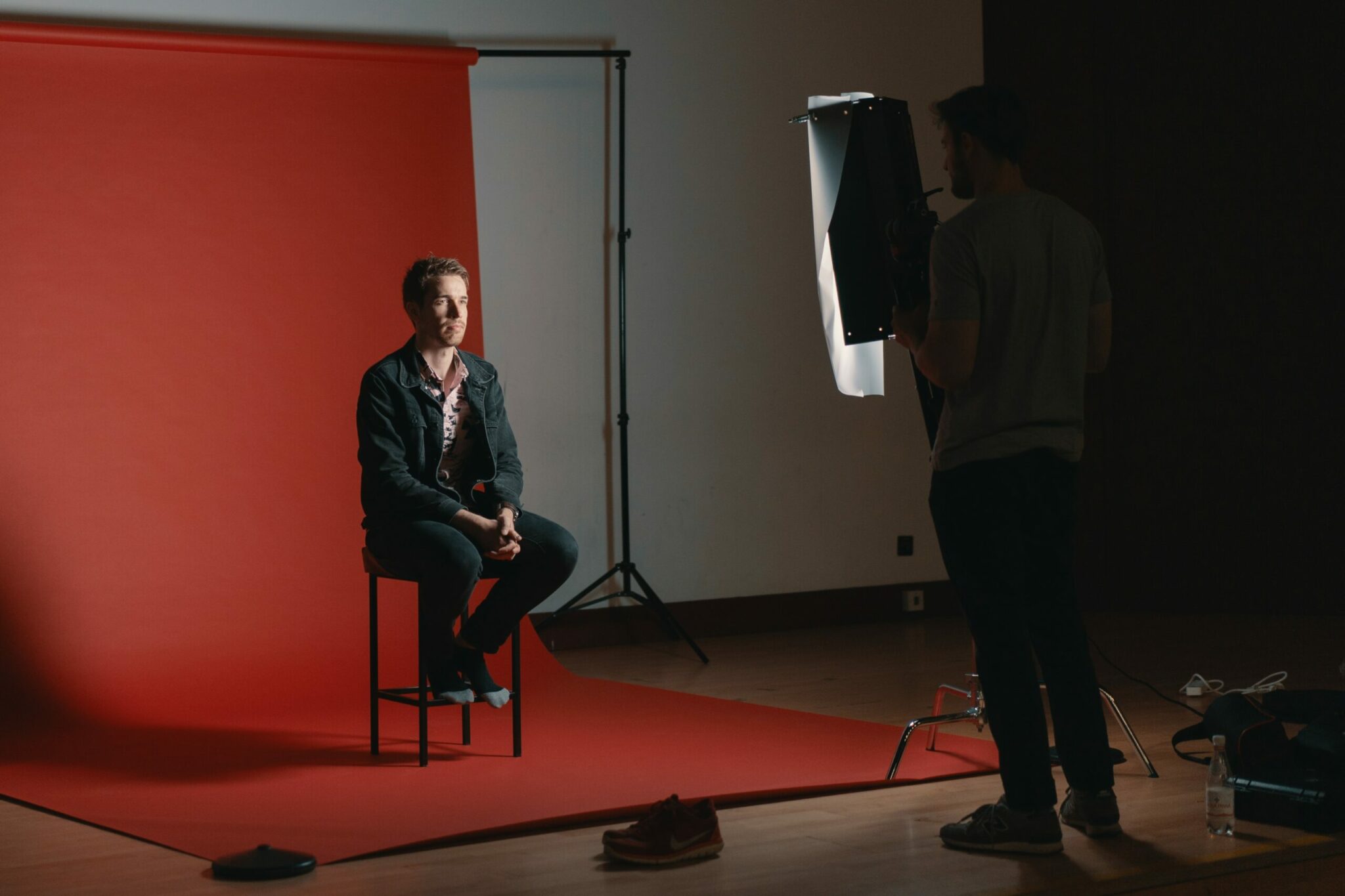 Un homme est assis sur un tabouret face à l'appareil photo sur un fond rouge. L'éclairage de studio et le photographe sont visibles dans cette configuration de studio photo.