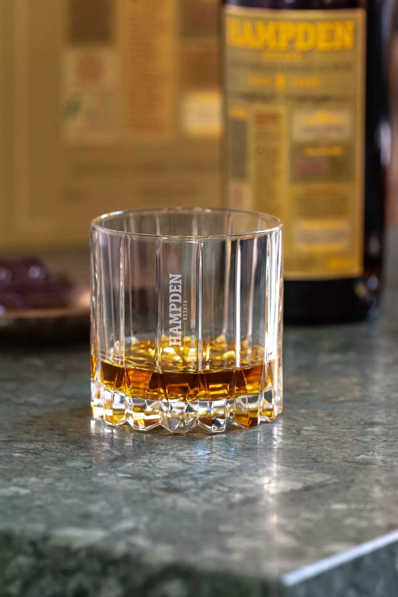 hampden-verrewebp Un verre de marque Hampden avec un liquide ambré sur un marbre vert ; bouteille floue avec une étiquette jaune en arrière-plan, éclairage de studio.