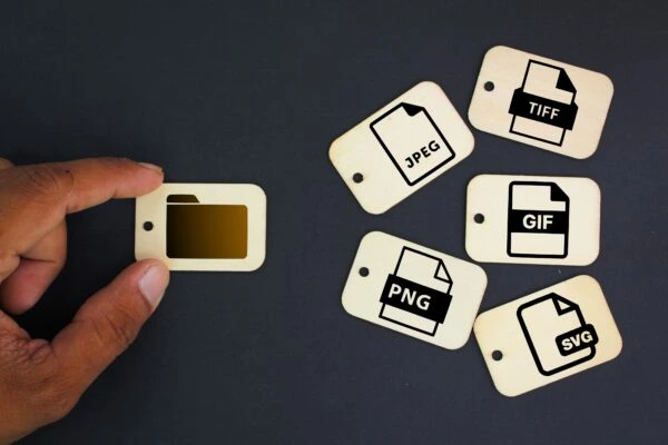 Une main présente une étiquette en bois avec une icône de dossier à côté d'étiquettes de formats de fichiers photo - JPEG, TIFF, GIF, PNG, SVG - sur un fond sombre.