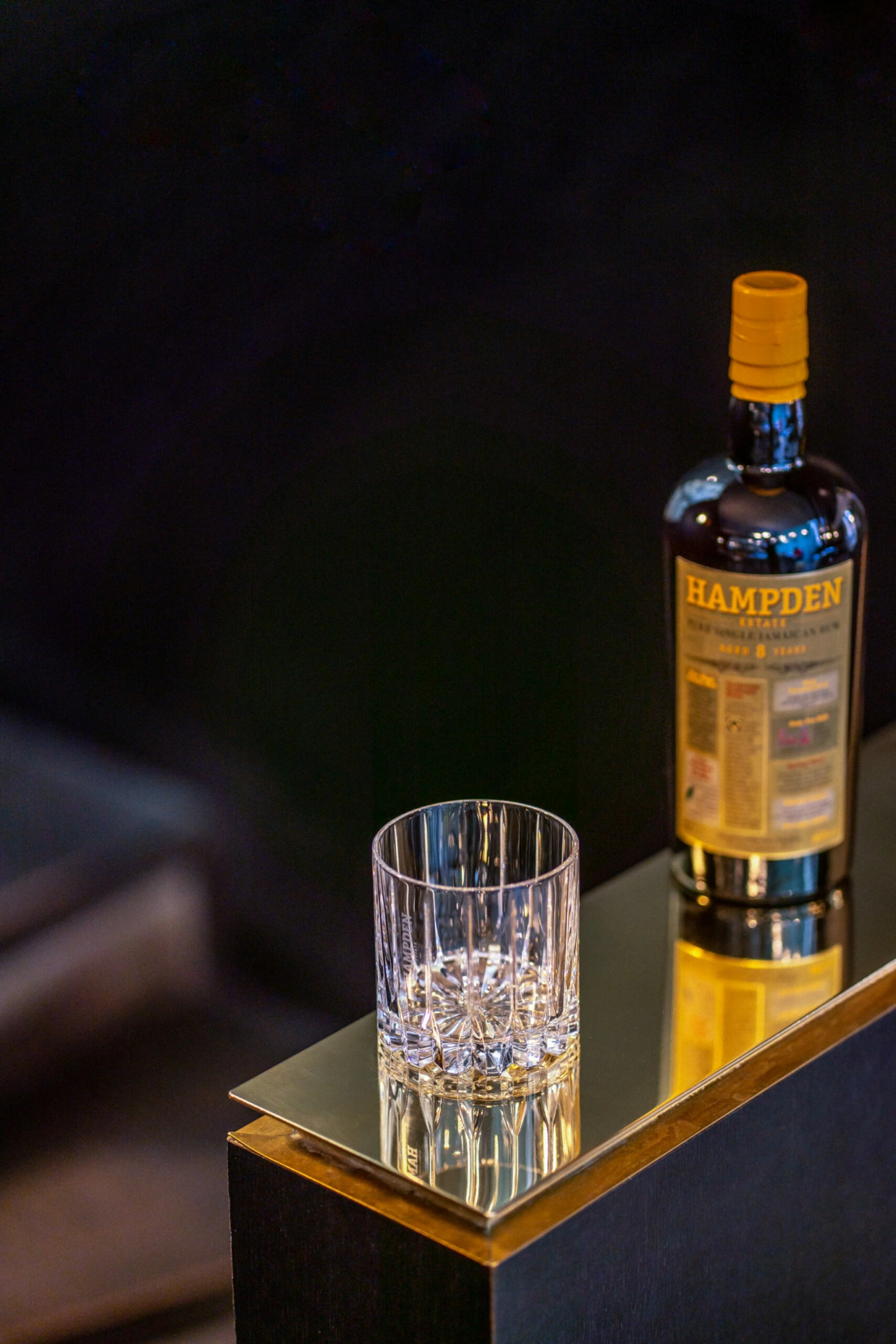 Packshot Hampden Verre + Bouteille Un verre transparent avec des glaçons sur une surface réfléchissante à côté d'une bouteille Hampden avec un bouchon jaune, capturé dans une configuration de photographie de produits.