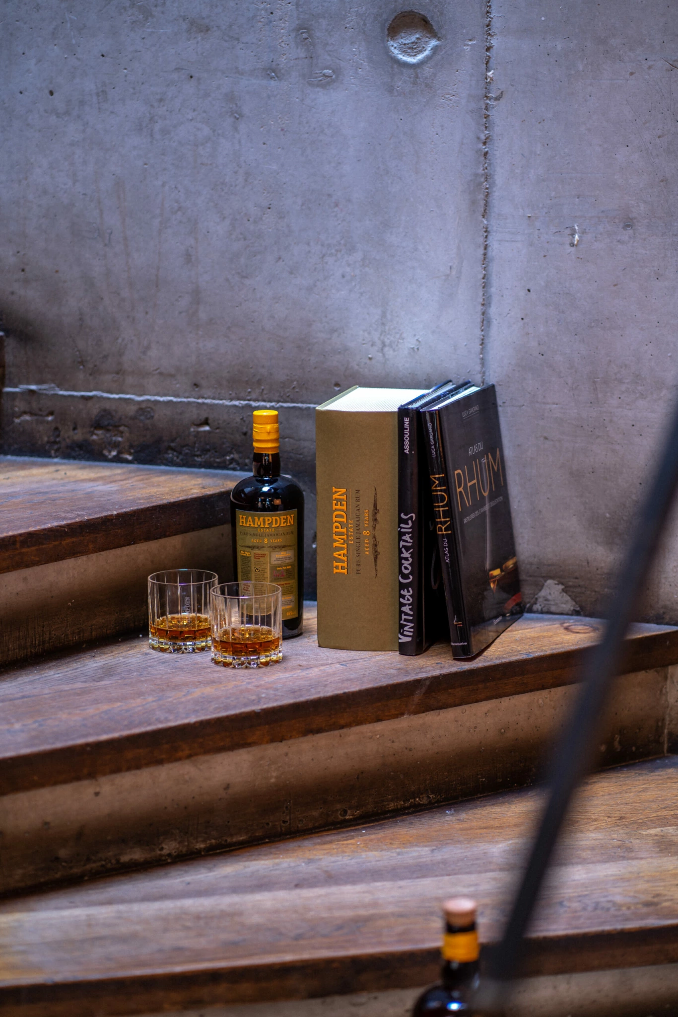 Photographie Ambiance Hampden Escalier Une bouteille de rhum Hampden, deux verres à whisky et des livres intitulés RHUM et HAMPDEN sont disposés sur des escaliers en bois près d'un mur en béton pour la photographie de produits.