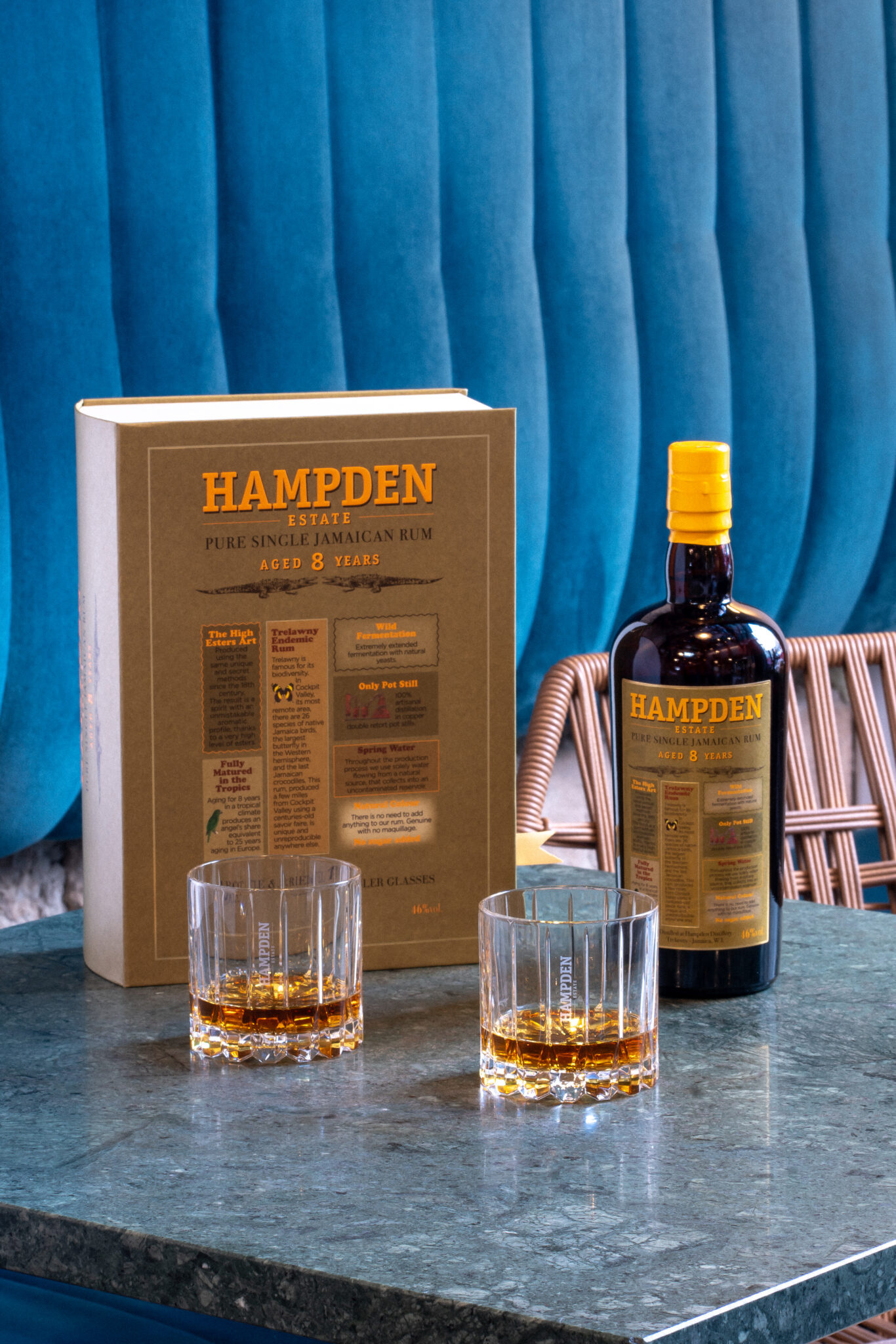 Photographie Ambiance Hampden sur une Table Rhum jamaïcain 8 ans d'âge Hampden Estate, boîte et deux verres versés disposés sur une table en marbre vert avec un fond tapissé bleu.