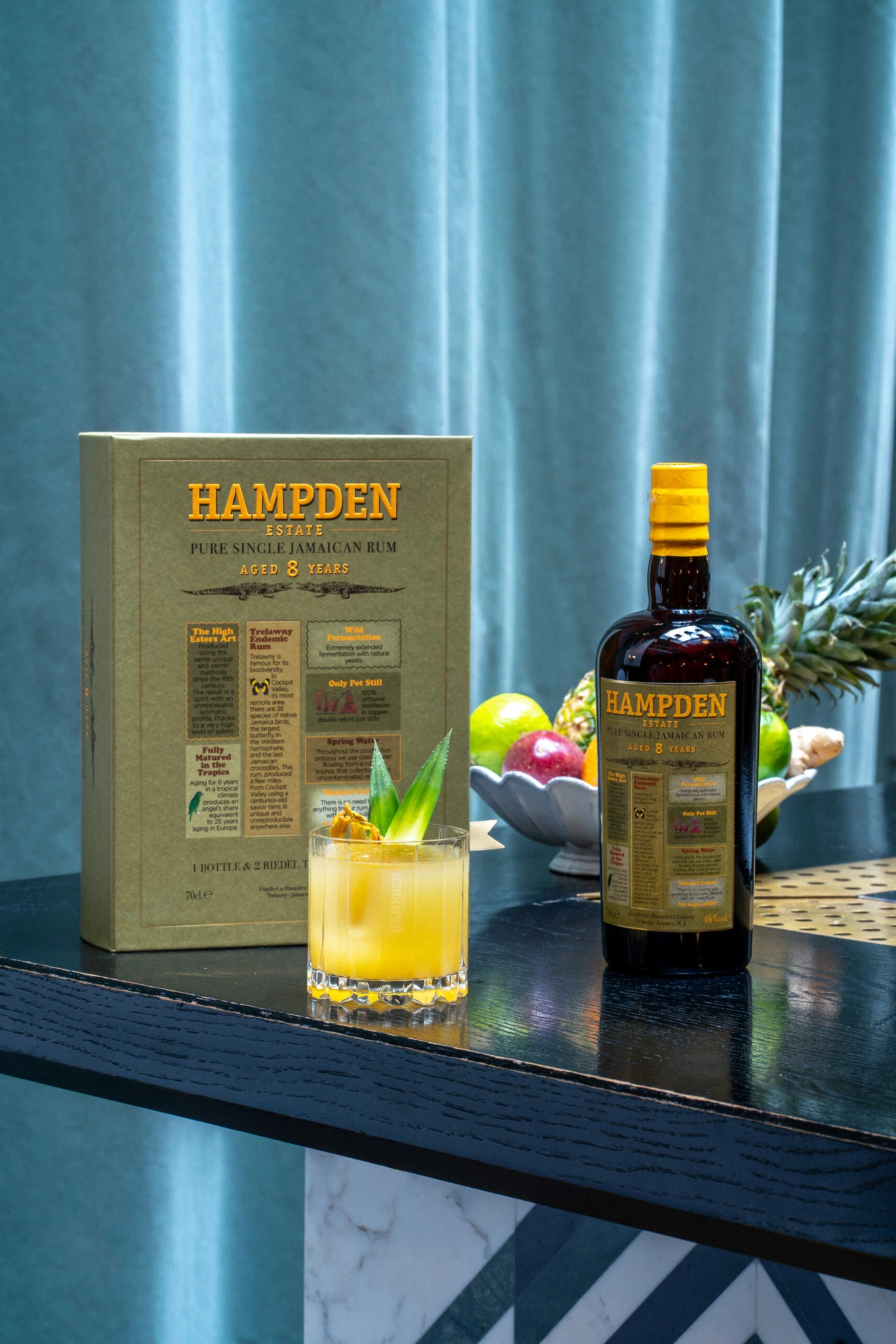 Photographie Ambiance Hampden sur une Table Cocktail Une bouteille de rhum vieux Hampden Estate avec son coffret à côté d'un cocktail verni à l'ananas sur un comptoir, des fruits et des rideaux en arrière-plan.