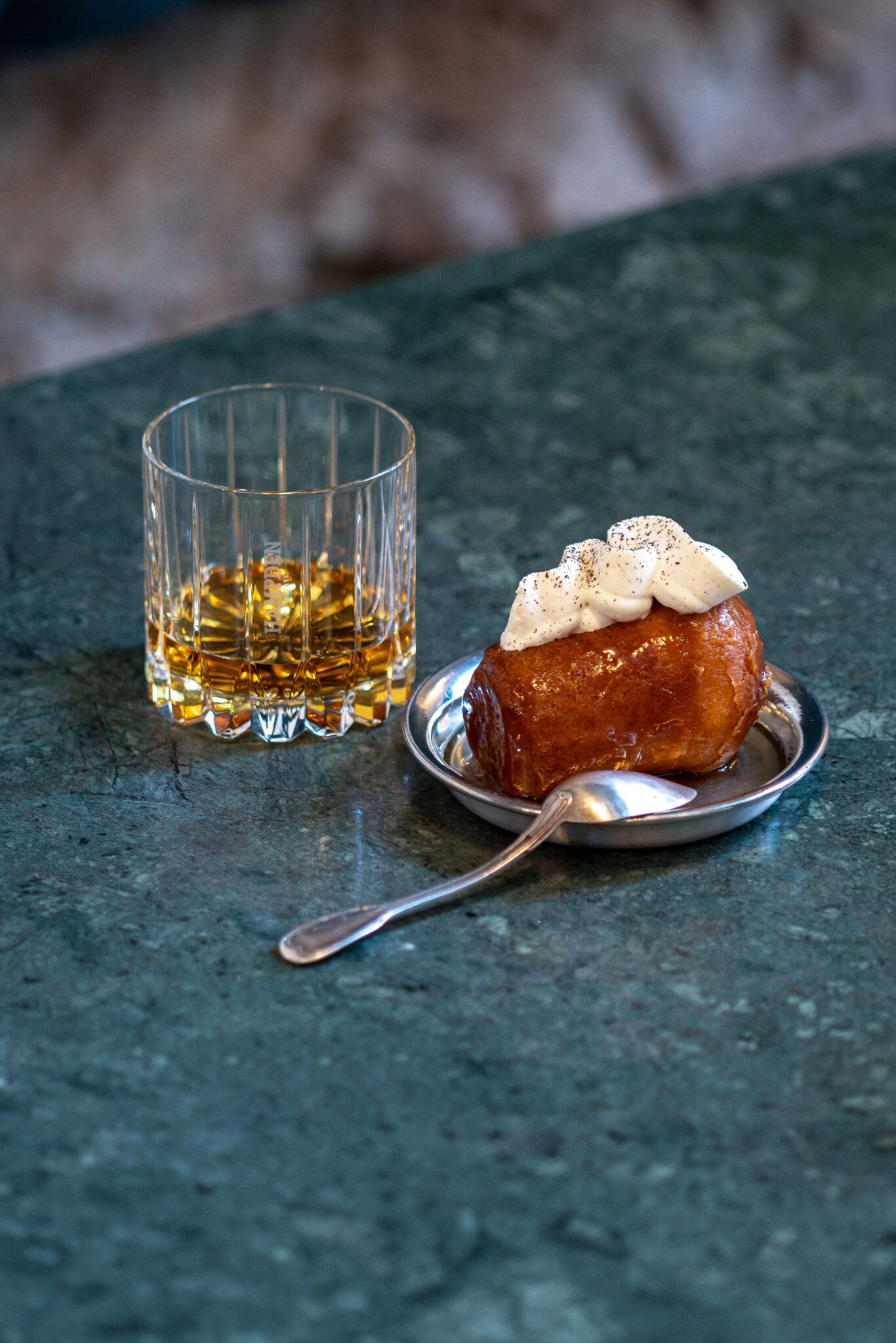 Photographie Ambiance Hampden sur une Table Verre + Baba au Rhum Verre de liqueur ambrée et plat en métal avec de la crème fouettée sur du marbre vert, la cuillère à côté - idéal pour la photographie commerciale de produits alimentaires.
