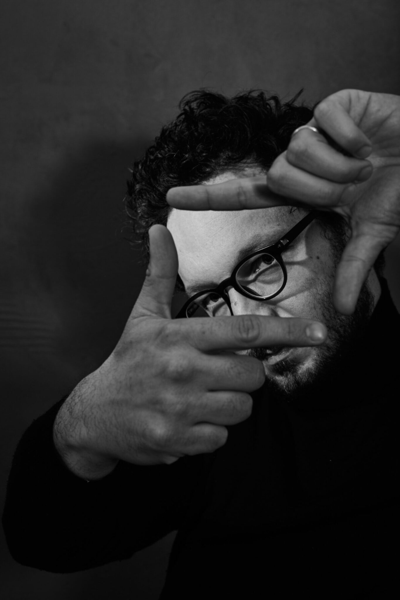 Un individu aux cheveux courts et bouclés et aux lunettes crée un cadre de main devant son visage. Portrait en noir et blanc.
