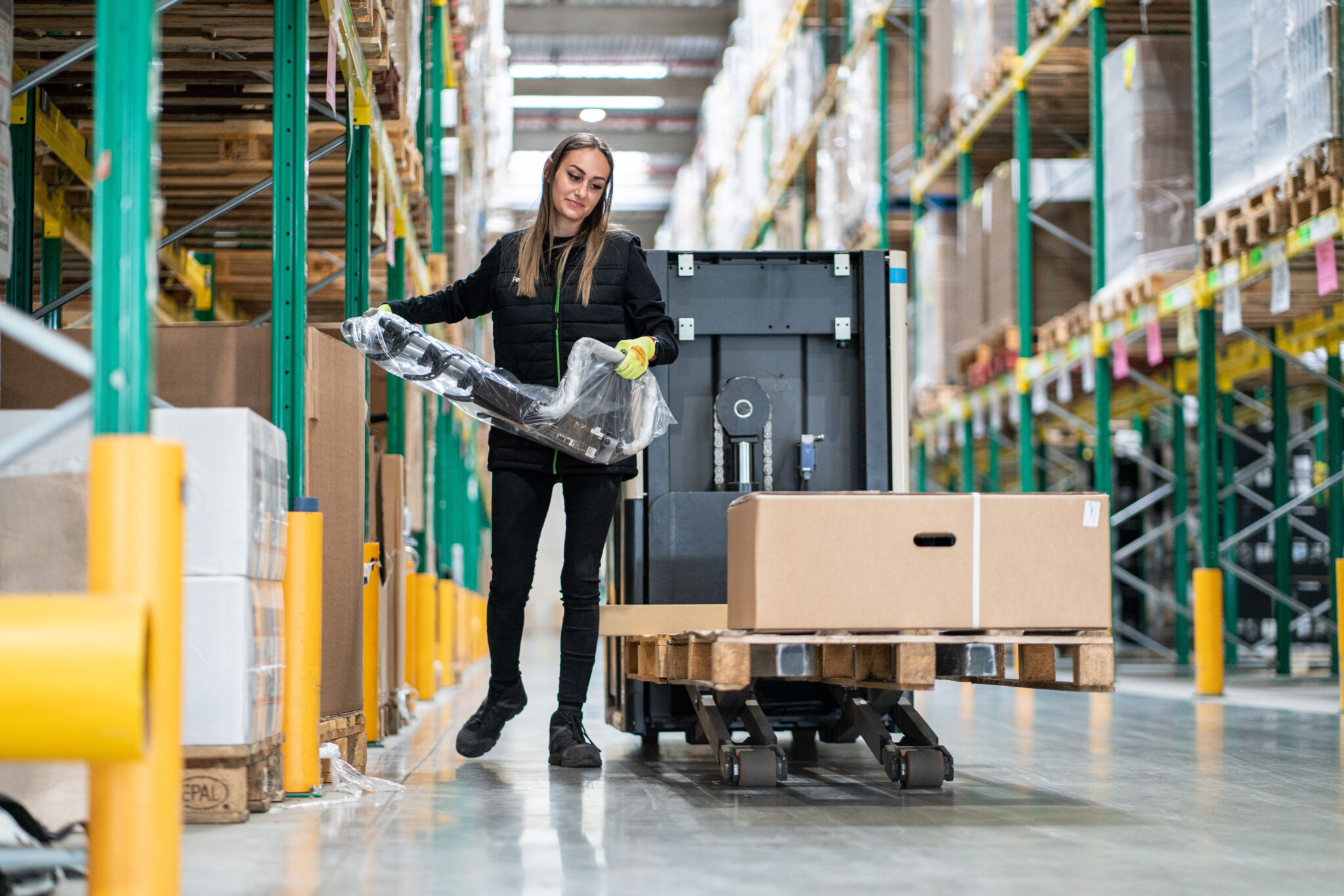 Reportage Photo Corporate Logistique Feu Vert 10 Une femme vêtue d'une veste noire et de gants déballe le plastique d'une palette de boîtes dans l'allée d'un entrepôt, capturé dans un style documentaire candide.