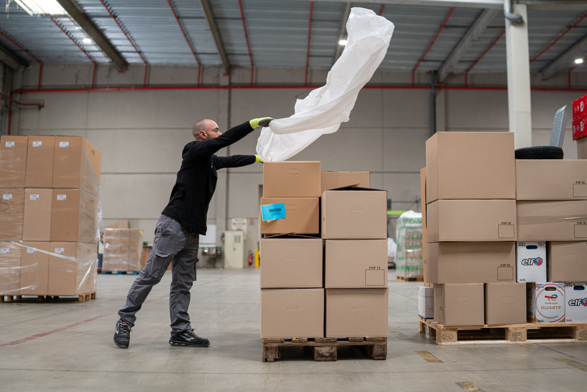 Reportage Photo Corporate Logistique Feu Vert 5 Un entrepôt à hauts plafonds dans le Rhône avec des sols en béton ; un ouvrier soulève le plastique des boîtes en carton, capturé dans un style documentaire.
