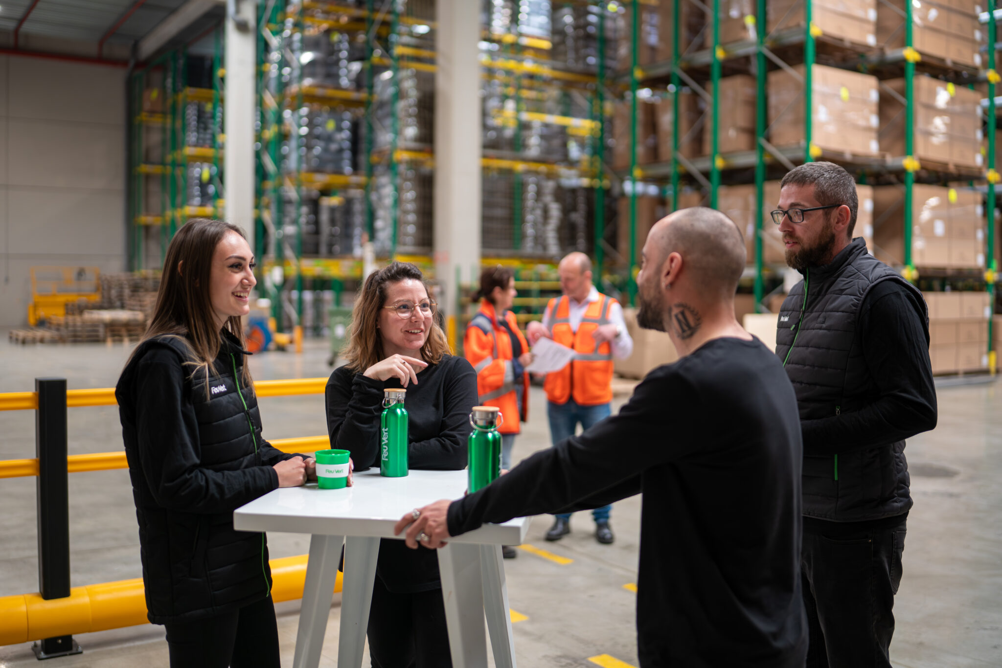 Reportage Photo Corporate Logistique Feu Vert 7 Quatre personnes se tiennent à une table haute dans un entrepôt du Rhône, parlant et tenant des bouteilles réutilisables ; des étagères et des travailleurs forment l'arrière-plan.