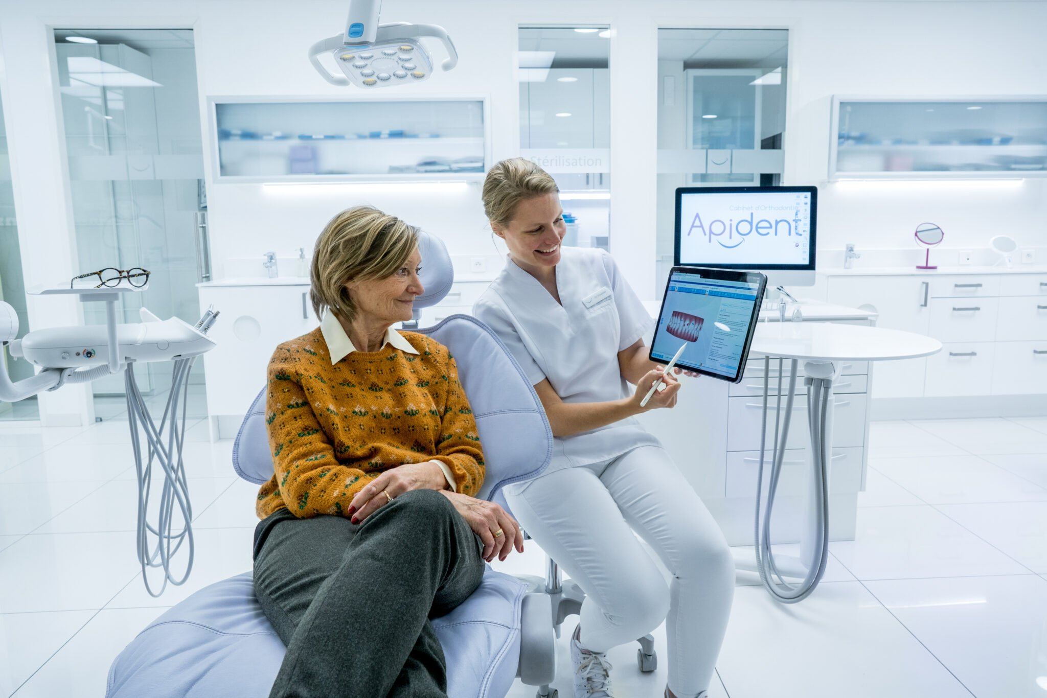 shooting Apident mise en scène 6 Orthodontiste présentant une image dentaire numérique à un patient dans une clinique contemporaine, avec équipement et moniteur Apident dans le cadre.