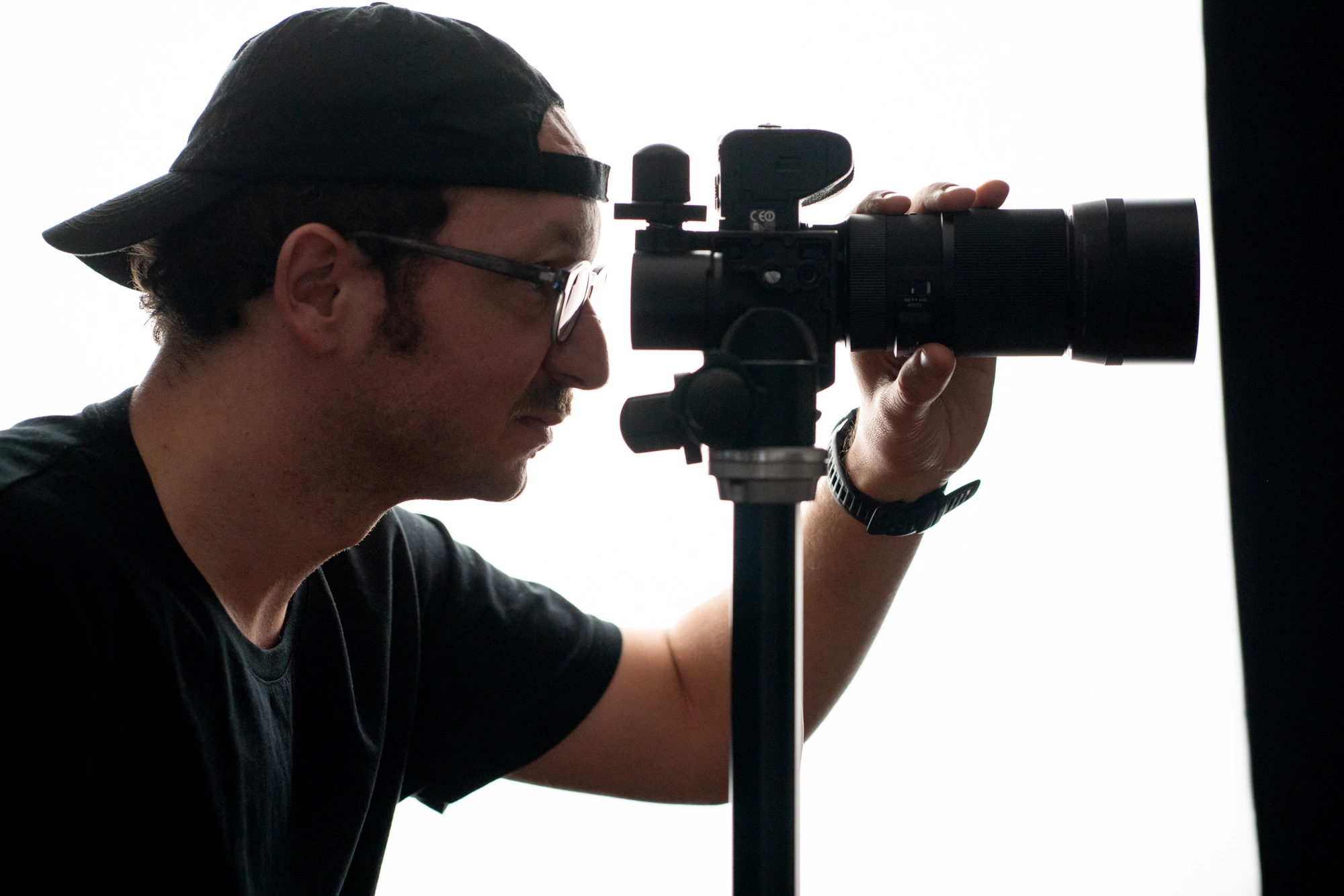 Un technicien de studio, vêtu d'une chemise noire et d'une casquette, étalonne un appareil photo reflex numérique sur un trépied, observant les réglages à travers le viseur.
