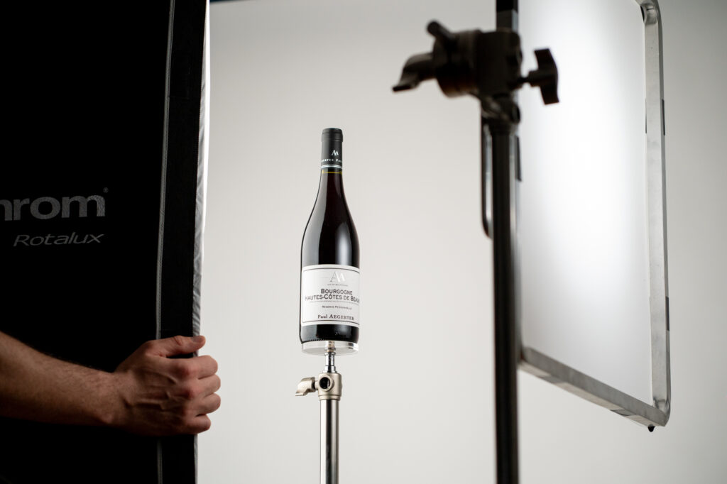 Bouteille de vin placée sur un support de studio avec du matériel d'éclairage ; la main d'un technicien ajuste la boîte à lumière sur un fond blanc uni.