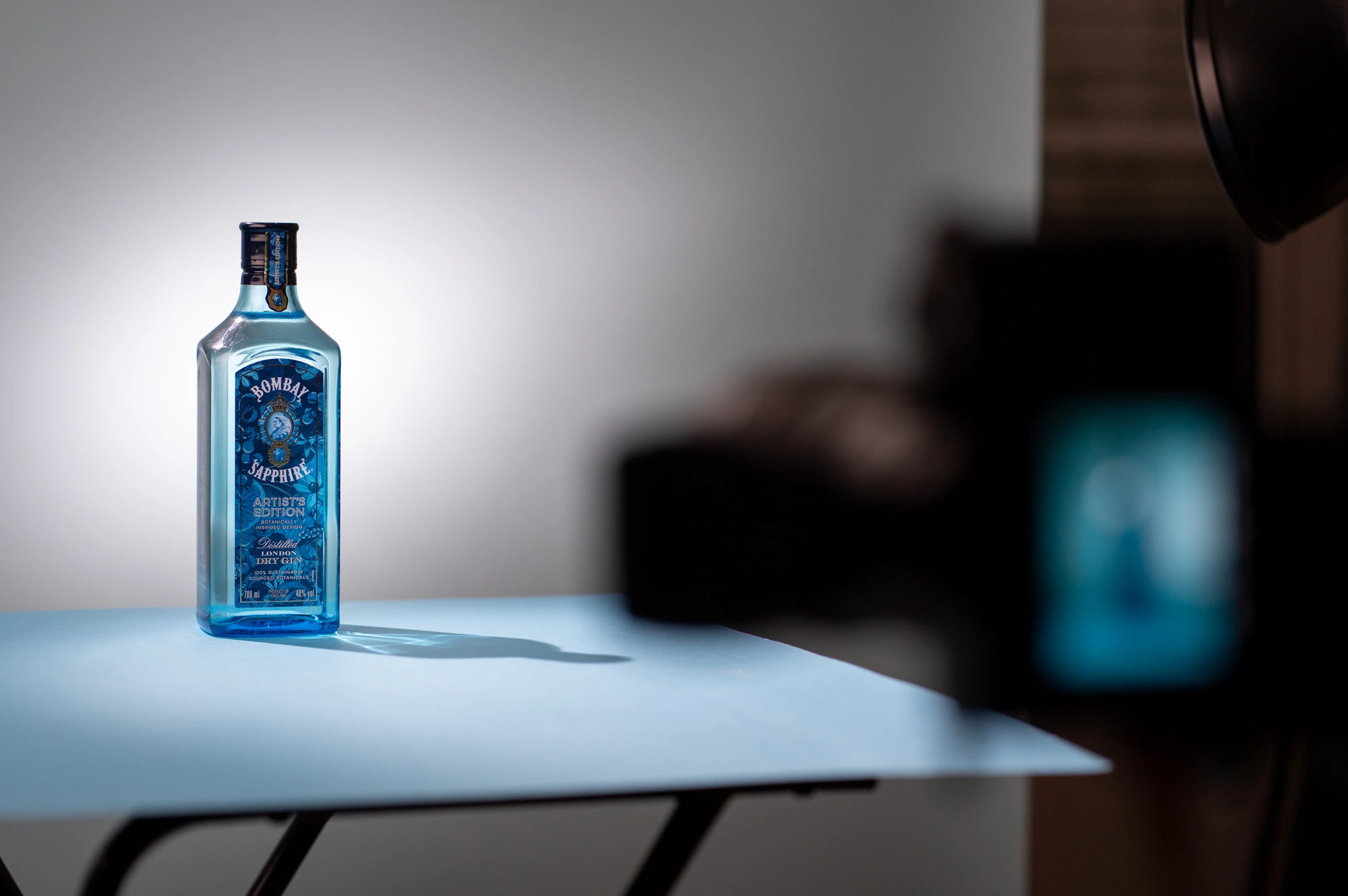 Une bouteille de gin Bombay Sapphire Artists Edition est placée sur une surface bleu clair avec un appareil photo flou au premier plan pour une photographie de produit.