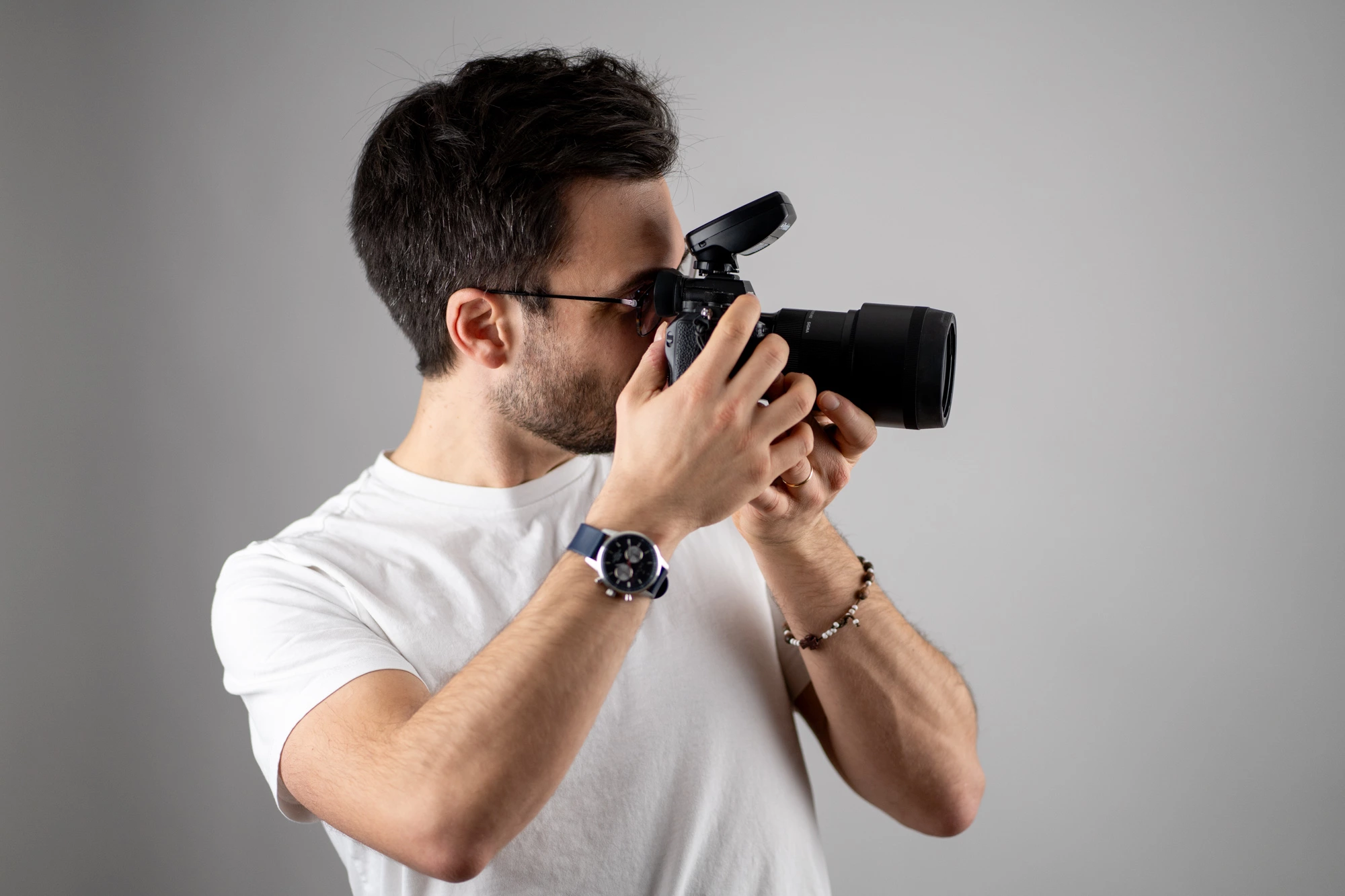 Un photographe professionnel vêtu d'un t-shirt blanc et d'une montre-bracelet utilise un appareil photo reflex numérique et prend des photos sur une toile de fond de studio grise.
