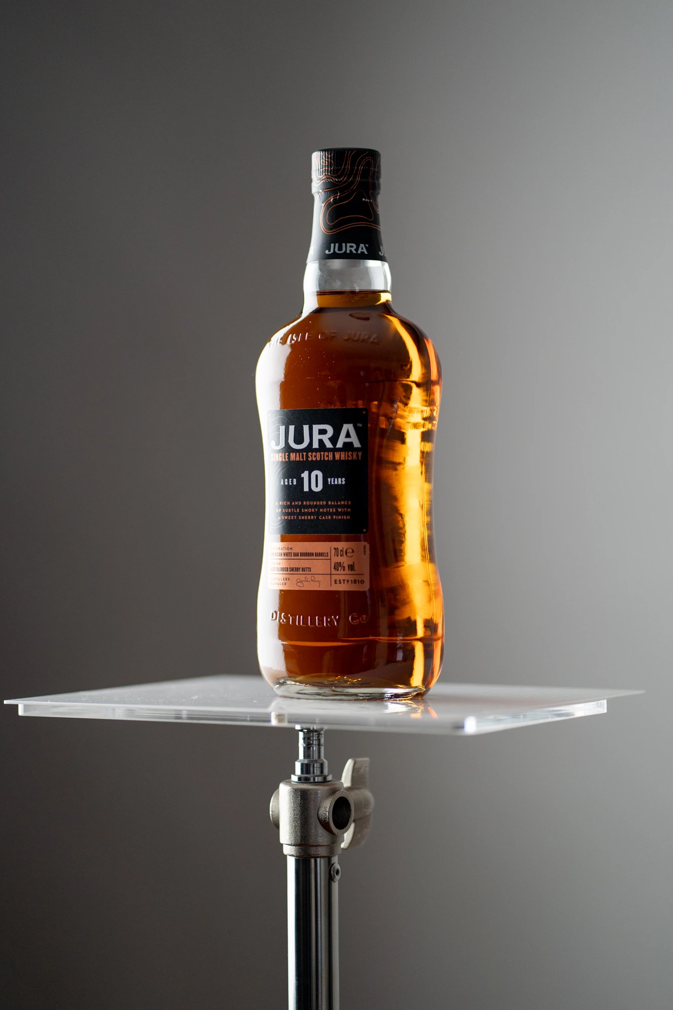 Bouteille de whisky écossais Jura 10 ans d'âge positionnée sur une surface acrylique avec un arrière-plan gris uniforme, adapté à la photographie de produits.