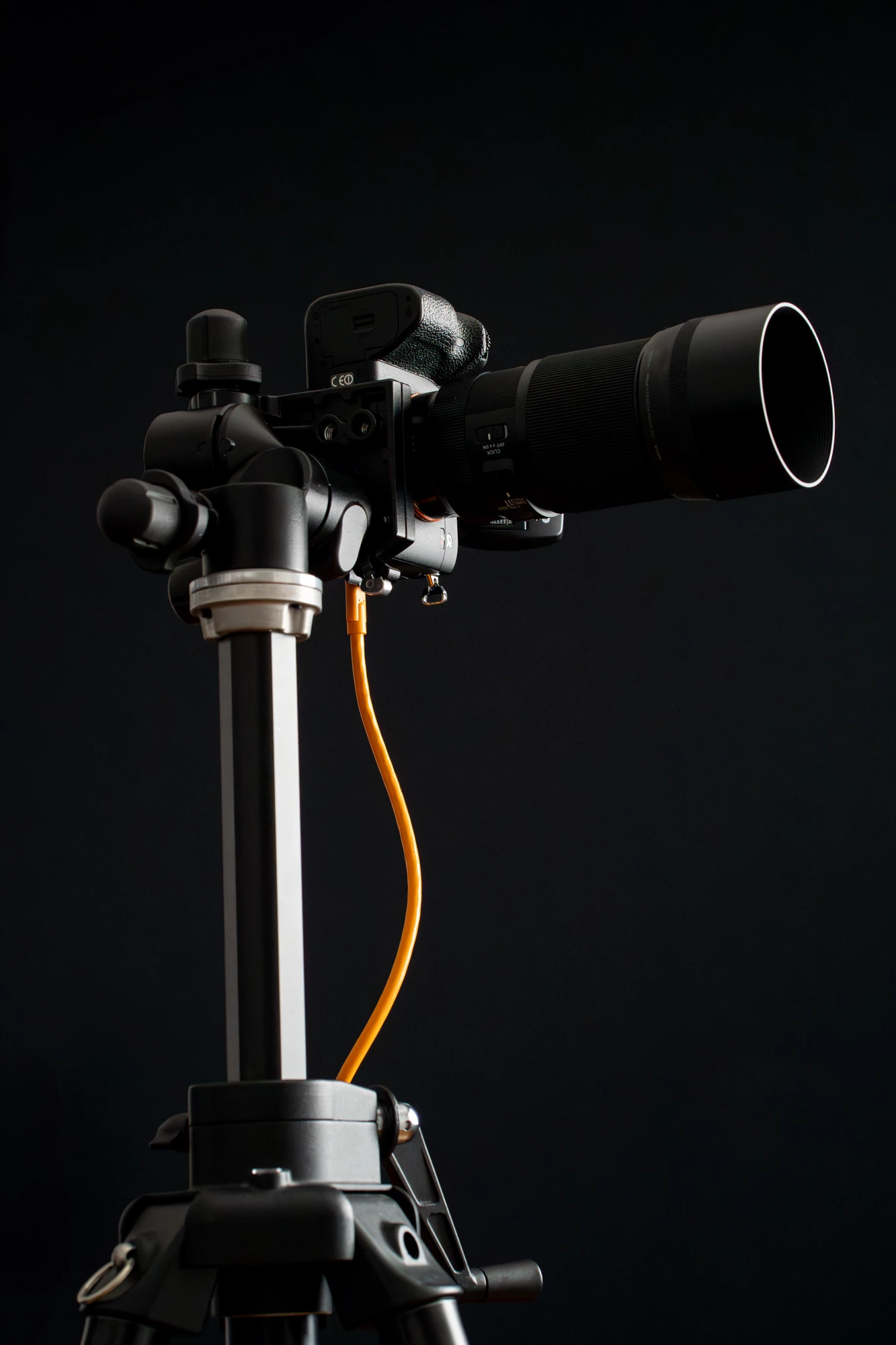 Un appareil photo reflex numérique équipé d'un téléobjectif est fixé sur un trépied de studio à l'aide d'un câble d'attache orange, sur une toile de fond noire unie.