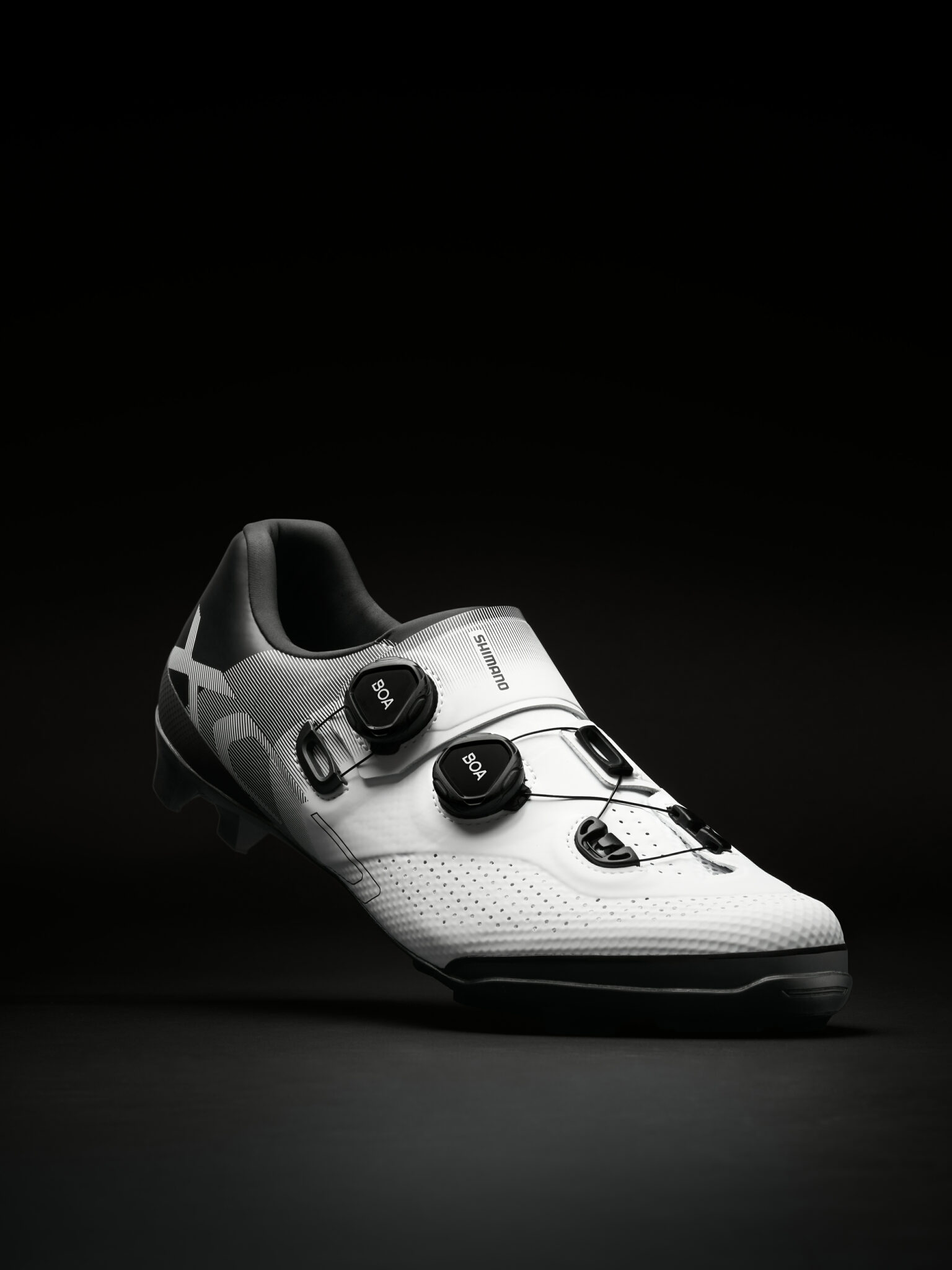 SHIMANO XC07 HD Une chaussure de cyclisme blanche et noire avec deux cadrans BOA et une tige perforée est éclairée en studio sur un fond sombre, mettant en évidence les détails techniques.