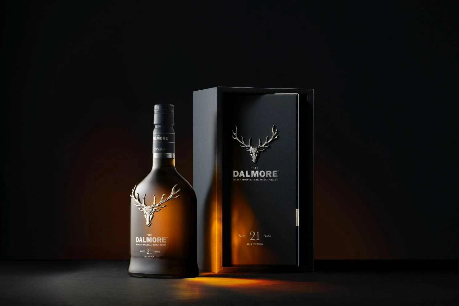 Bouteille de whisky Dalmore 21 ans d'âge et coffret noir avec logos de cerfs argentés présentés côte à côte sur un fond de studio sombre et bien éclairé.