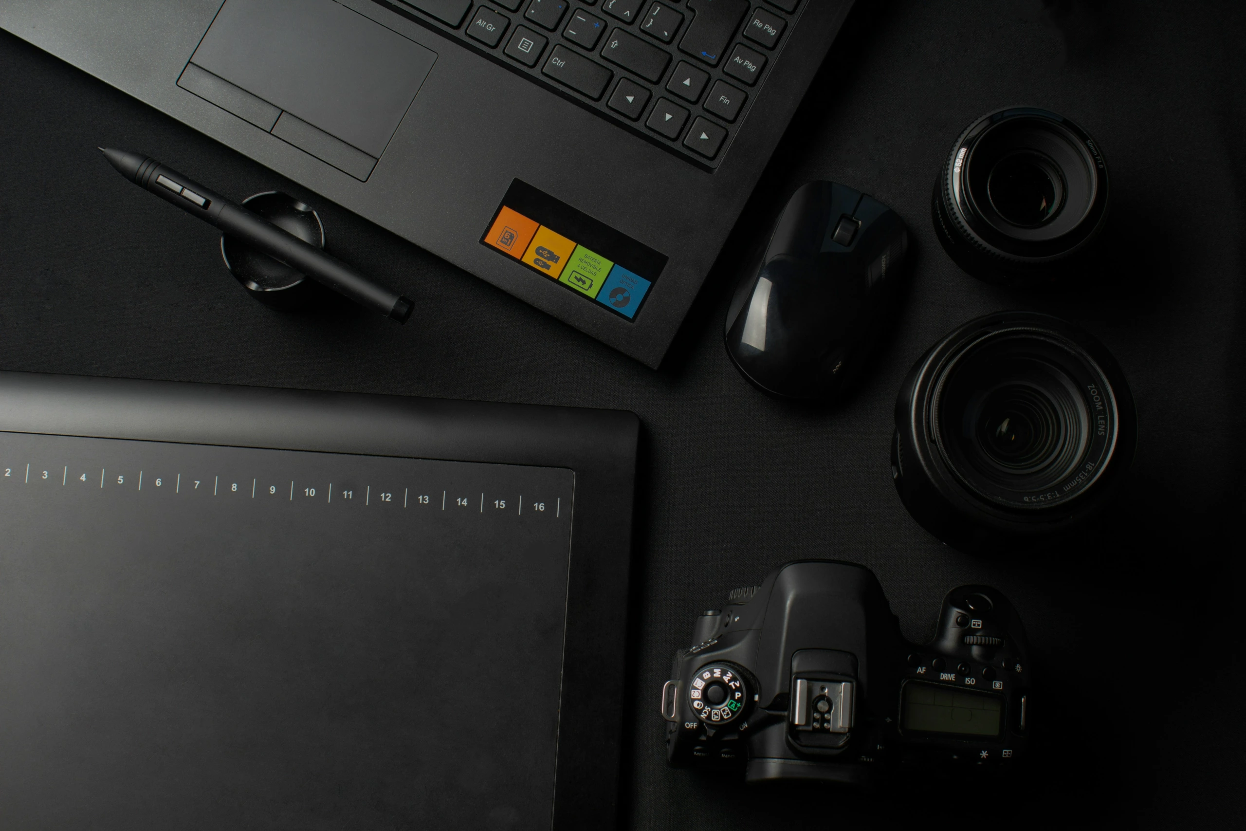 Vue à plat d'un ordinateur portable, d'une tablette graphique avec stylet, d'un appareil photo reflex numérique, de deux objectifs interchangeables et d'une souris sur un espace de travail noir.