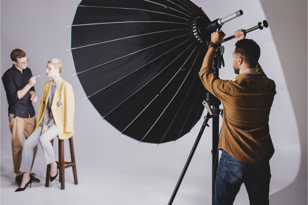 Dans un studio de photographie, un photographe ajuste l'éclairage tandis qu'une maquilleuse prépare un mannequin vêtu d'un blazer jaune et d'un pantalon gris.