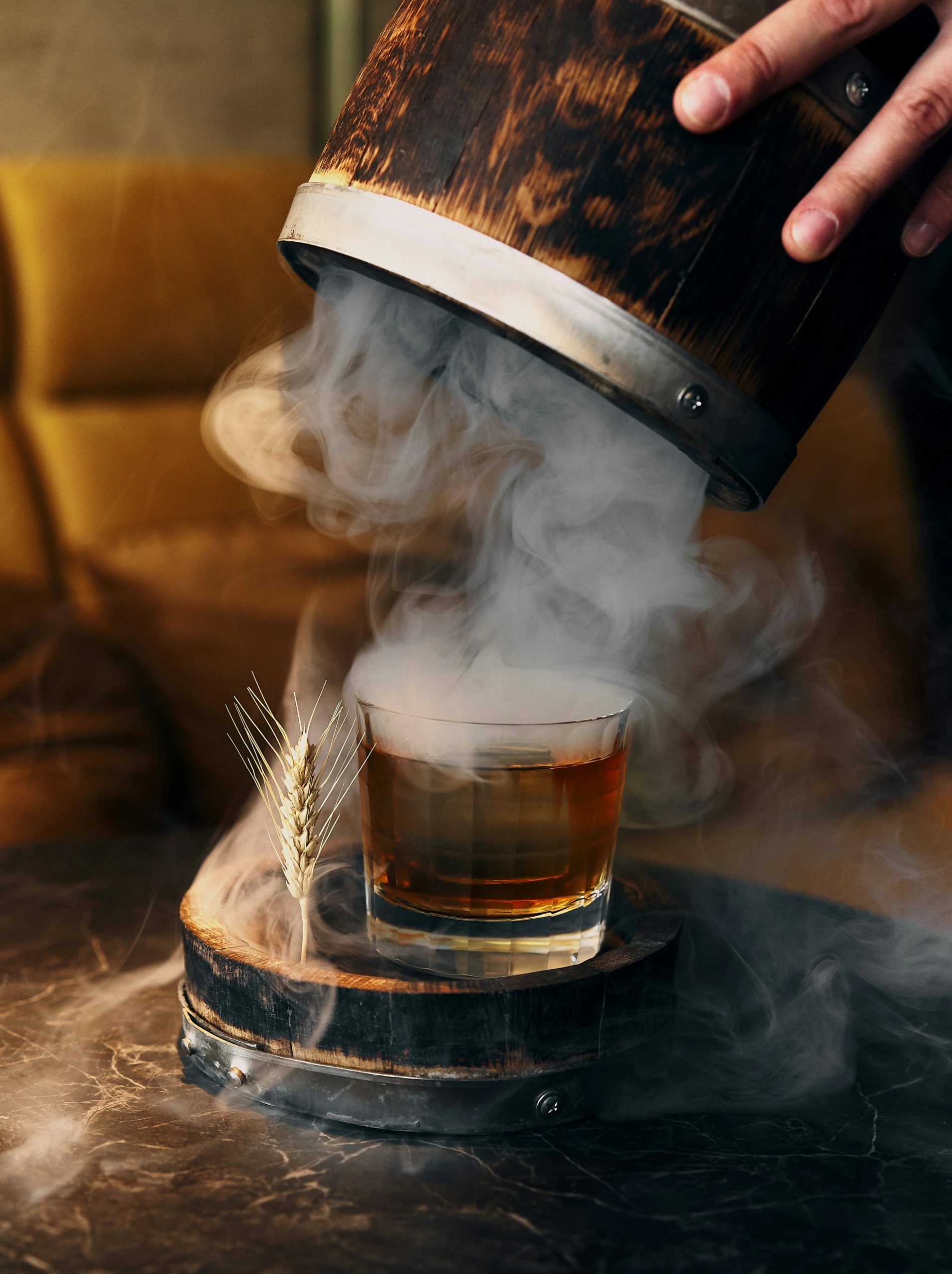 Un verre à whisky posé sur un sous-verre en bois émet de la fumée lorsqu'une main soulève le couvercle ; une garniture de blé repose à côté. L'éclairage de studio met en valeur les détails.