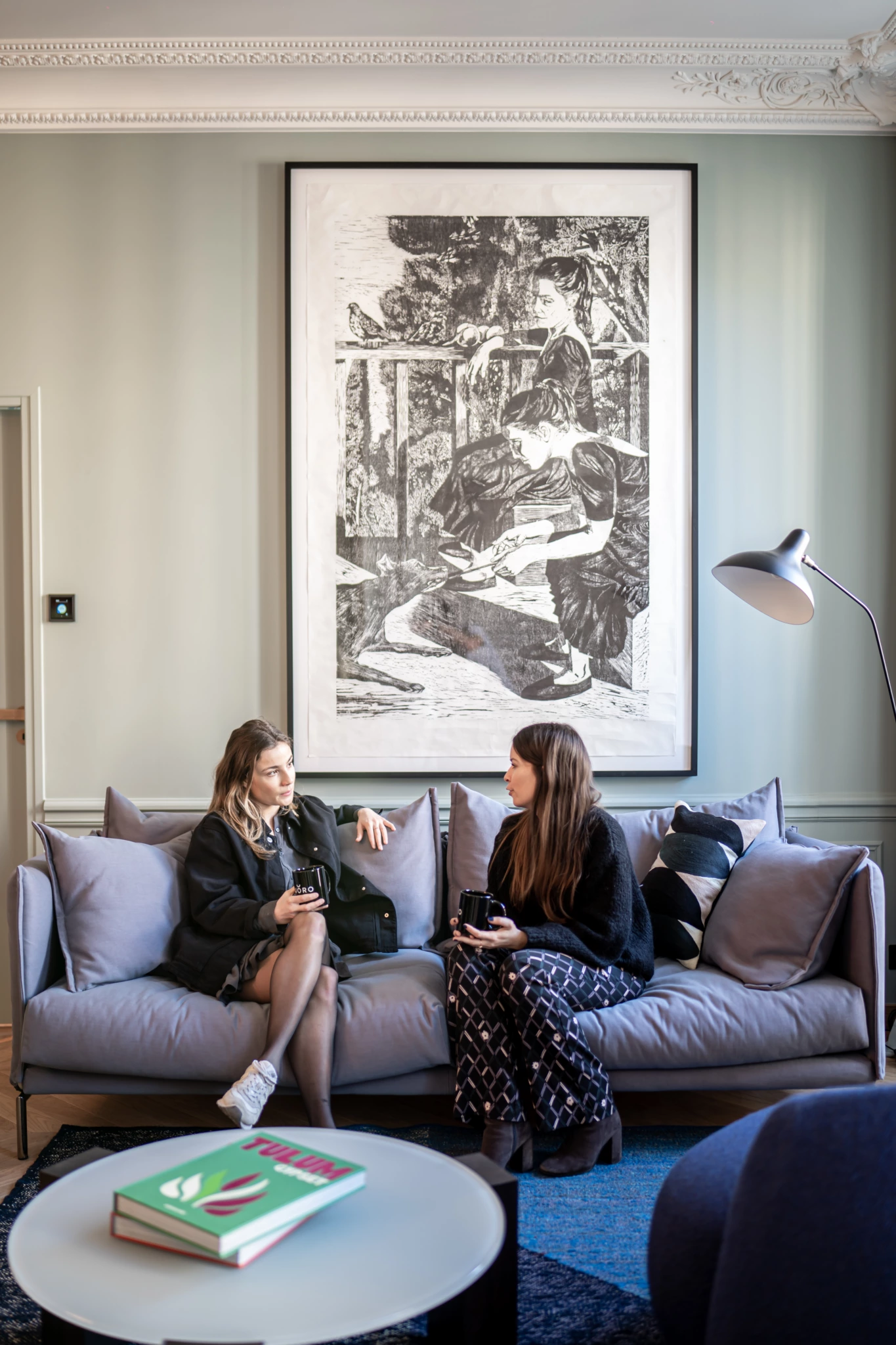 Mise en scene photo Joro 4 Deux femmes discutent sur un canapé gris avec des tasses pendant une séance de photos au bureau ; art en noir et blanc, lampe et livres visibles dans le cadre.