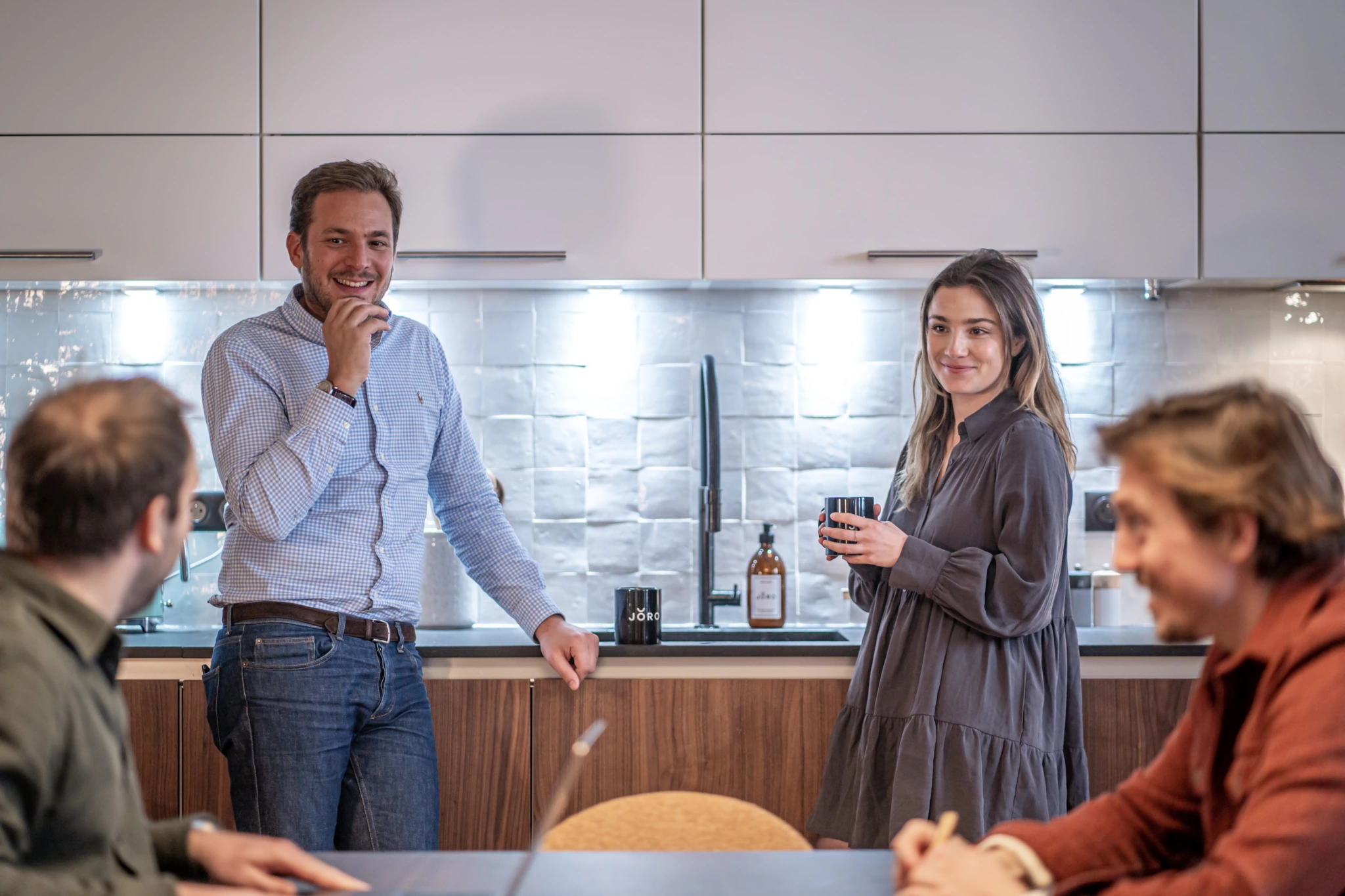 Mise en scene photo Joro 5 Photographie de bureau professionnelle de quatre collègues dans une cuisine moderne ; deux près de l'évier avec des tasses, deux conversant à la table.
