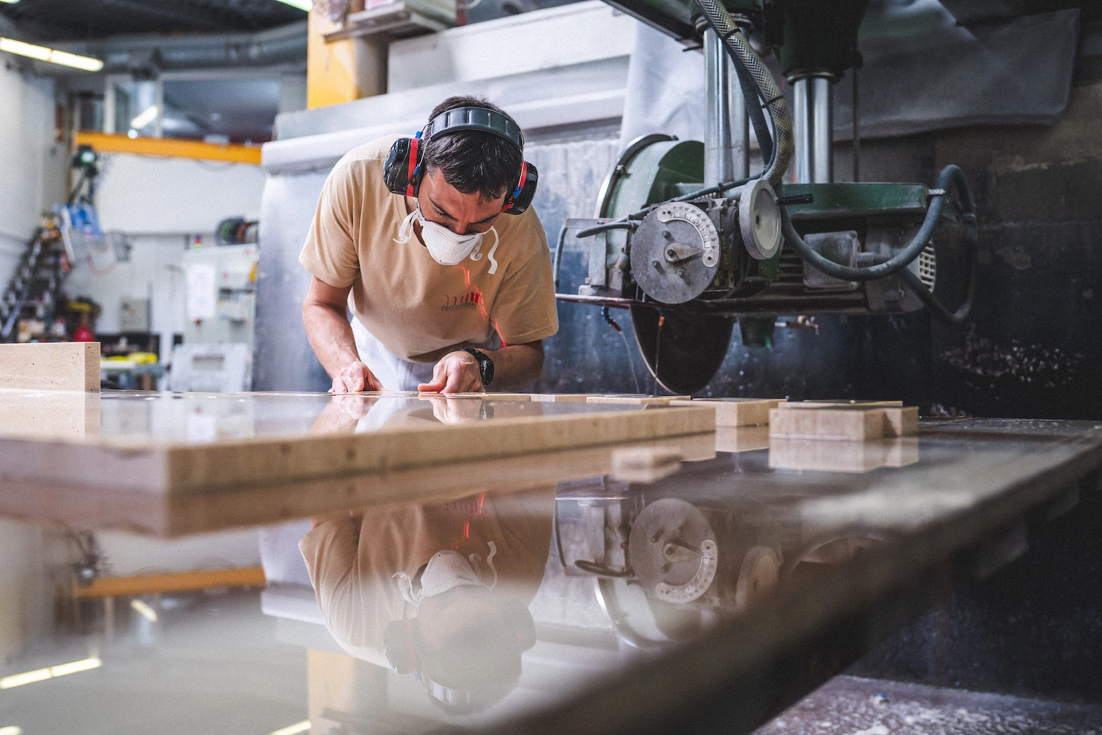 Un technicien muni d'une protection auditive et d'un masque examine une dalle de pierre dans un atelier ; machines industrielles visibles. Photographie de reportage d'entreprise.