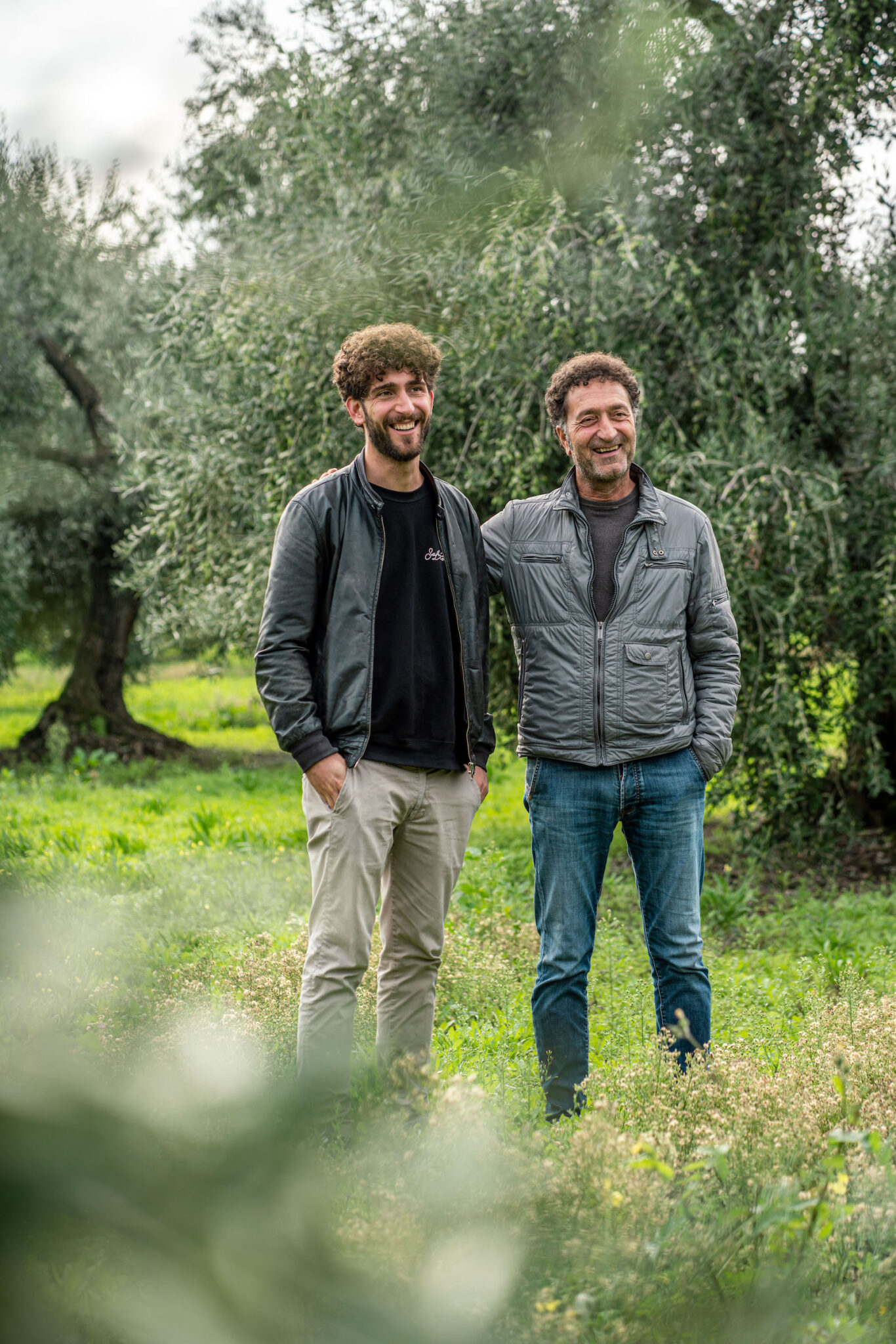 Portrait Metier Olivier & CO 13 Deux hommes vêtus de vestes et de pantalons décontractés posent en plein air dans une oliveraie herbeuse. Cette photo a été prise lors d'un reportage agricole d'entreprise.