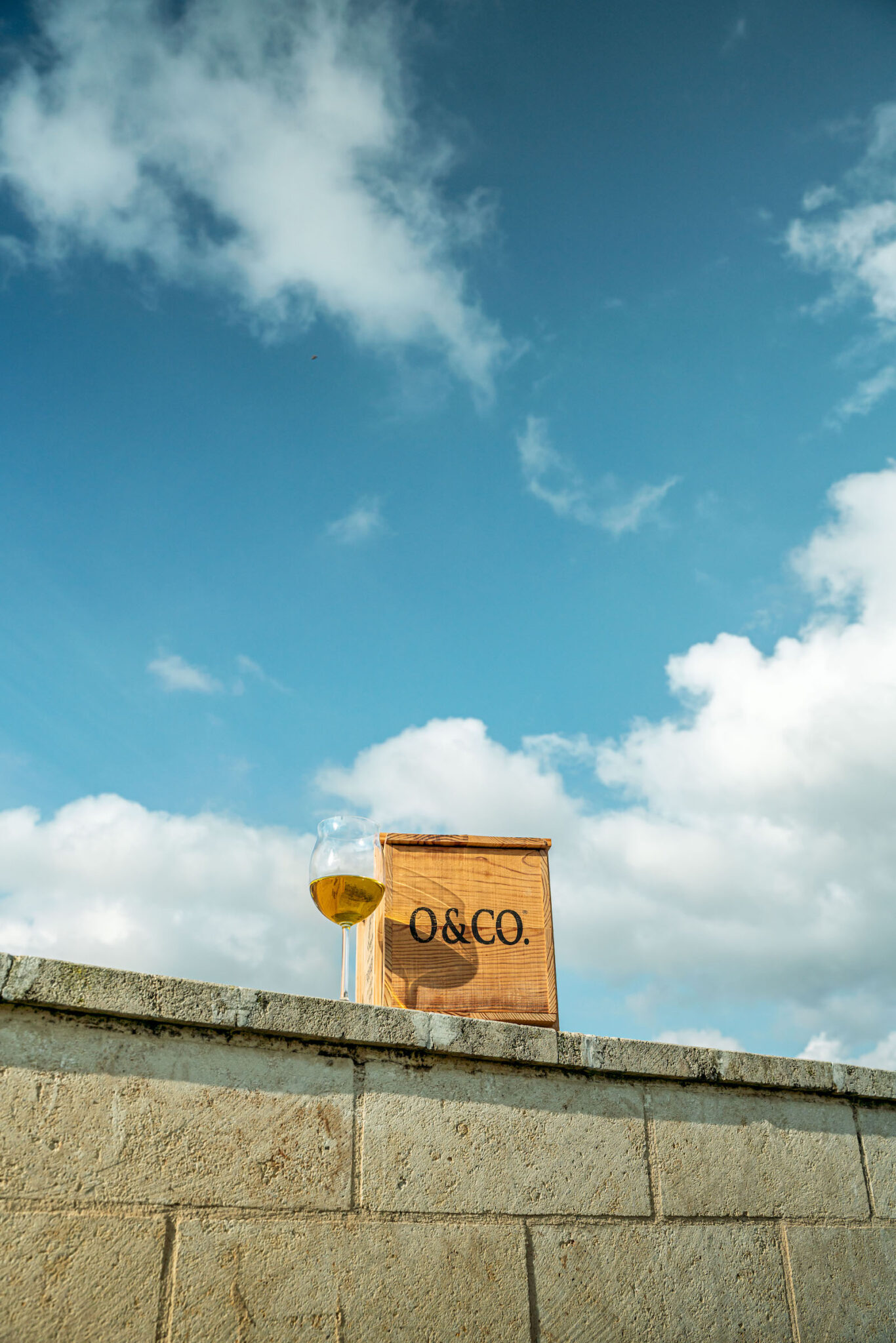 Portrait Metier Olivier & CO 19 Un verre de vin blanc et une boîte en bois O&CO. sur un mur de pierre sous un ciel bleu, photographiés pour un reportage agricole.