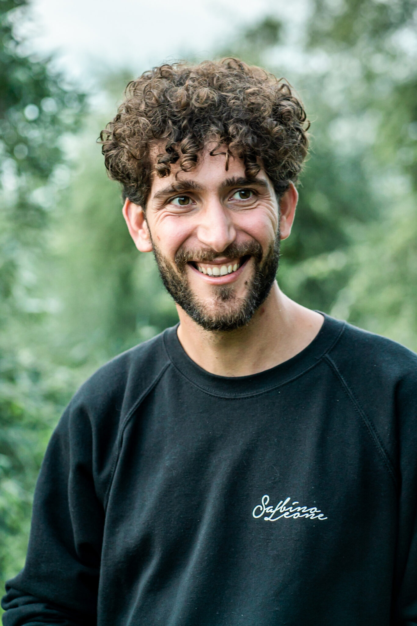 Portrait Metier Olivier & CO 8 Homme aux cheveux et à la barbe bouclés, en extérieur dans la verdure, vêtu d'un sweat-shirt noir ; photographie de reportage dans le domaine de l'agriculture d'entreprise.