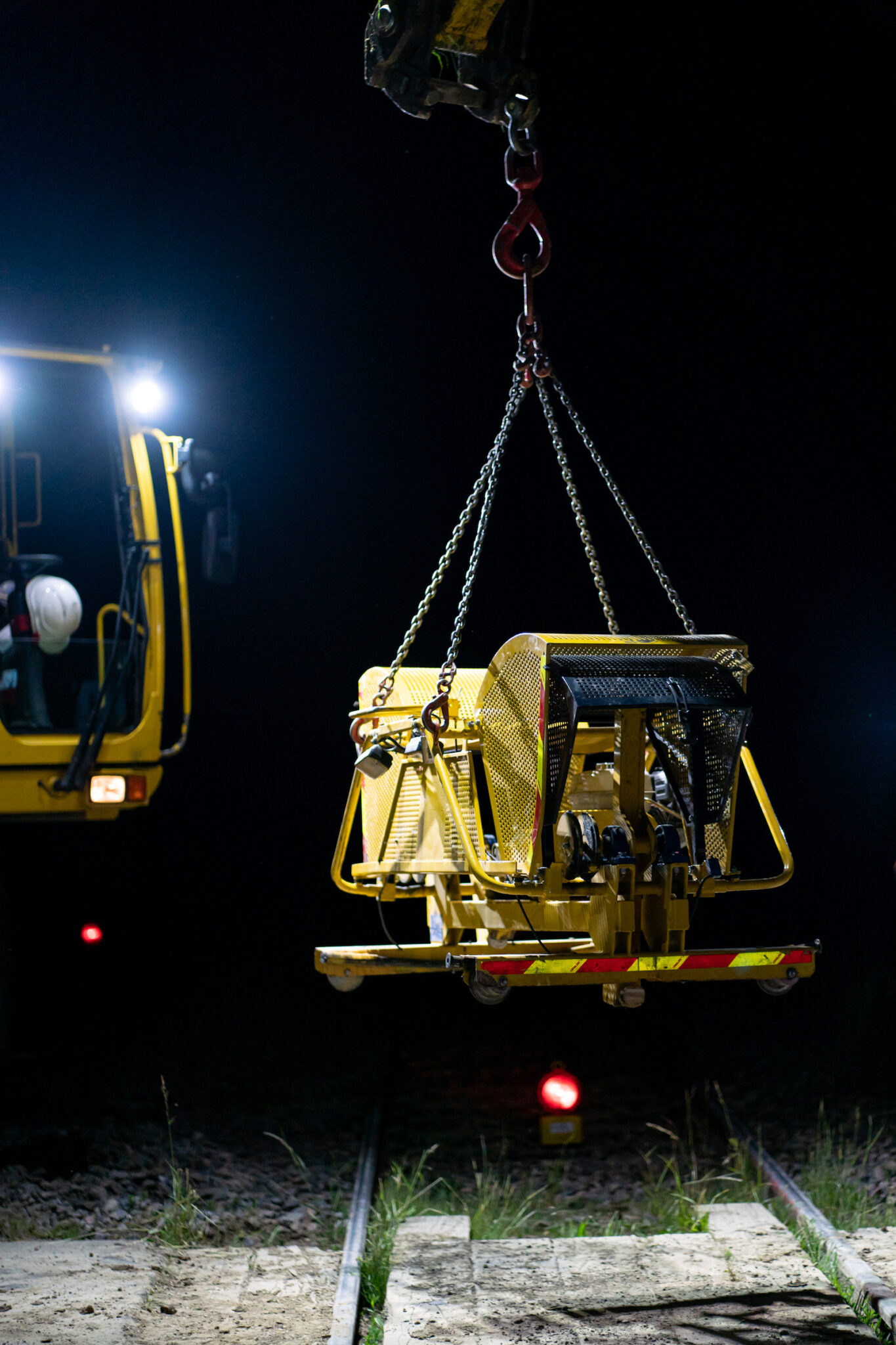 Portrait Metier Sferis 5 Photojournalisme nocturne d'une machine jaune hissée par une grue au-dessus d'une voie ferrée, avec une équipe dans un véhicule sous les faisceaux des phares.