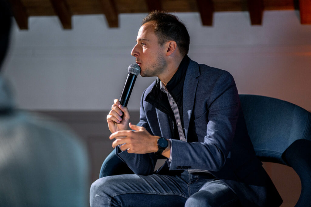 Photo de séminaire d'entreprise : homme en blazer bleu et jeans, assis, parlant au micro ; mur blanc, plafond en poutres de bois en arrière-plan.