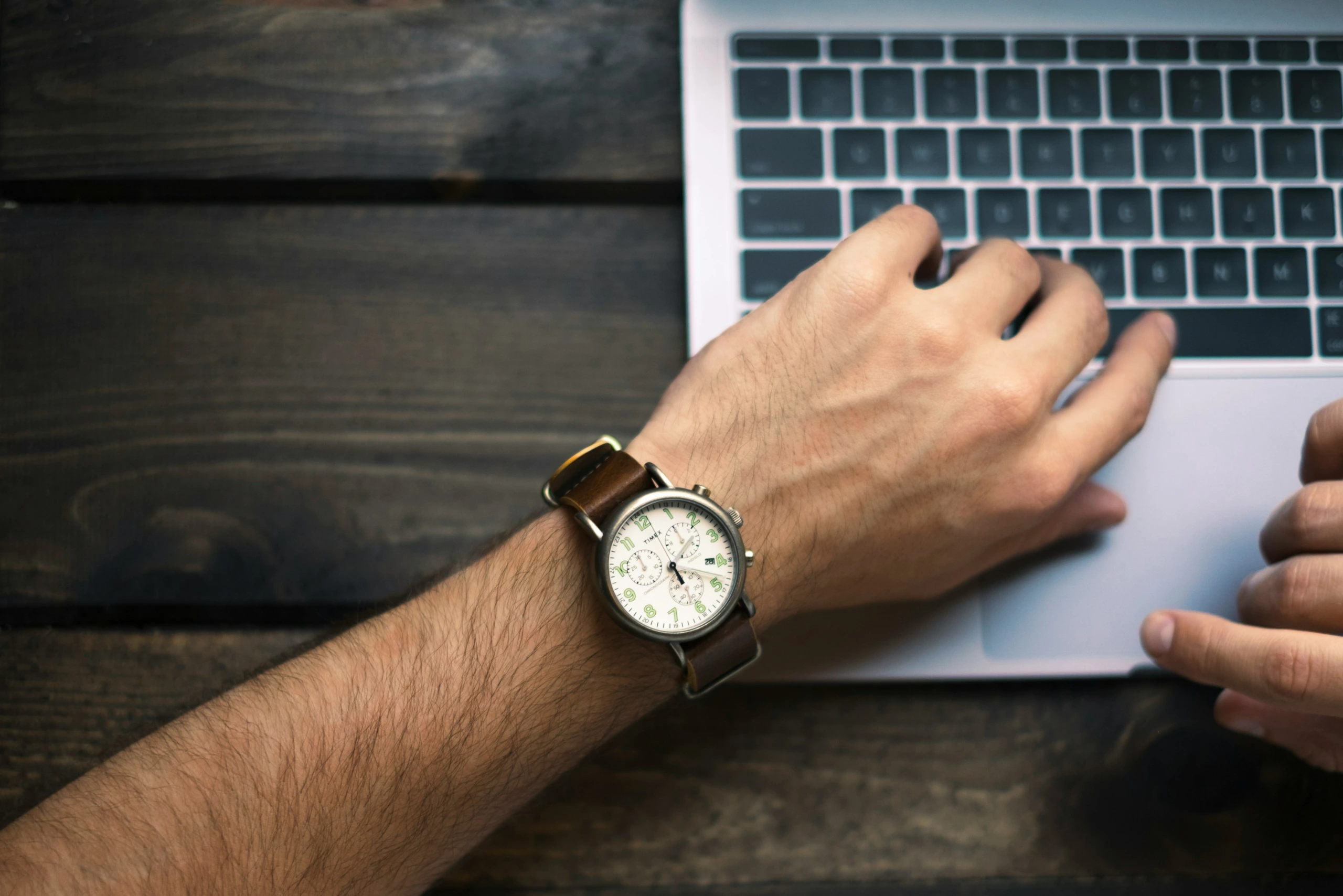 Individu avec montre-bracelet travaillant sur un ordinateur portable à un bureau en bois sombre, mains partiellement visibles, vue de dessus pour une composition d'espace de travail.