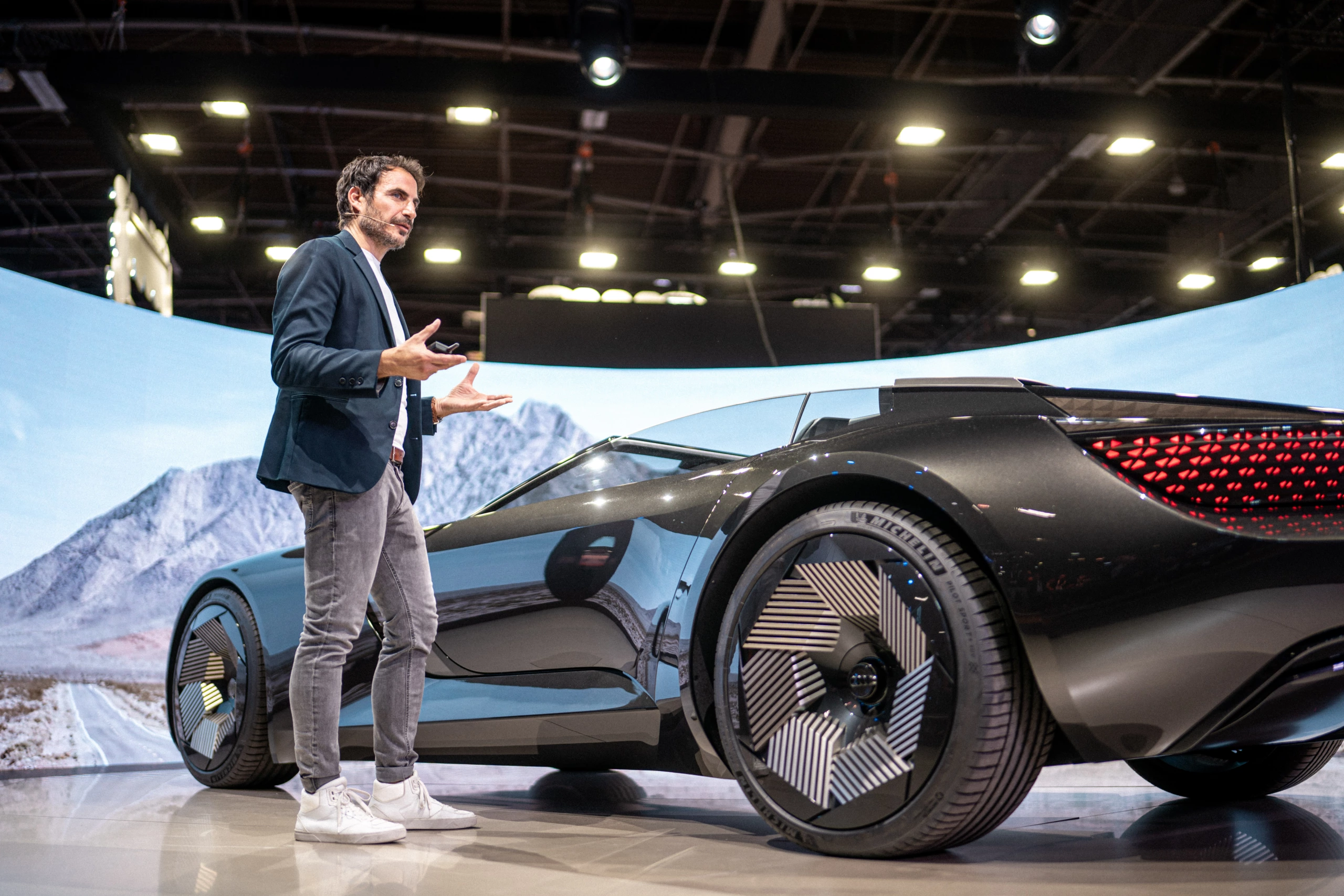 Un homme vêtu d'un blazer et d'un jean se tient à côté d'un concept car noir aux roues uniques lors d'un événement automobile en intérieur à Paris, avec une image en toile de fond.
