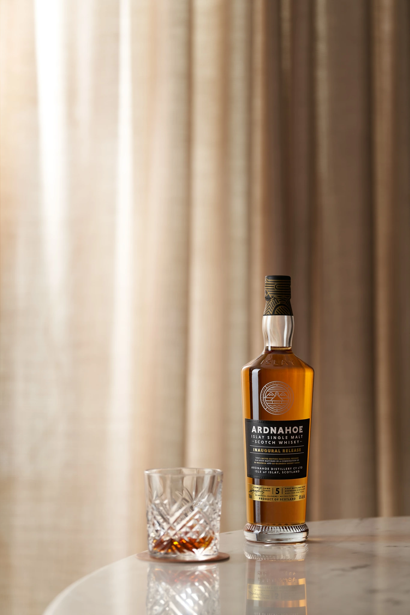 Ardnahoe Bouteille de whisky écossais Ardnahoe single malt et verre en cristal sur marbre, photographiés comme une nature morte avec des rideaux beiges en arrière-plan.