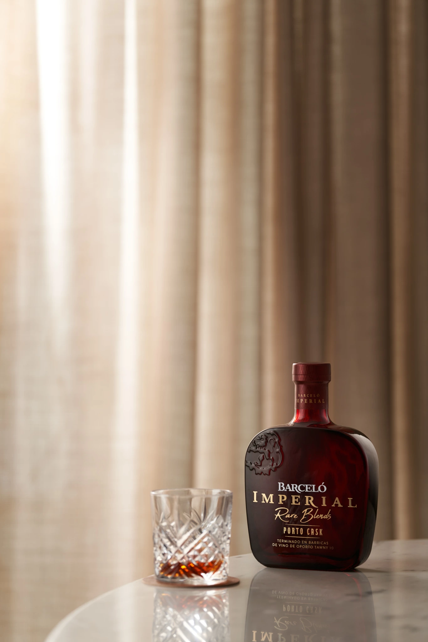 Barcelo Jona Un verre contenant un liquide ambré à côté d'une bouteille rouge foncé Barceló Imperial Ron Blended sur une surface blanche, mise en scène pour une photographie de produit.
