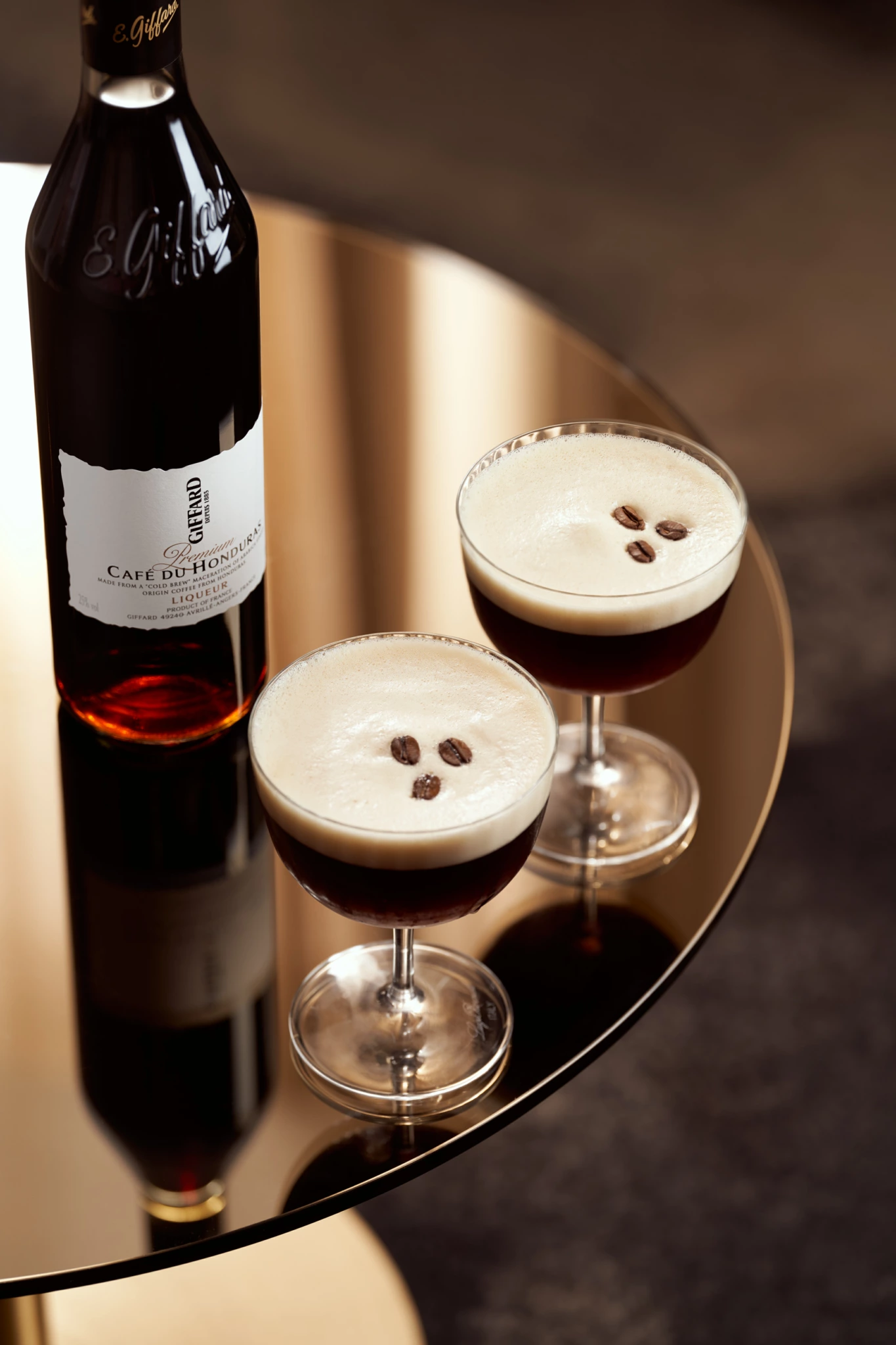 Giffard Café Bouteille de liqueur de café à côté de deux verres à coupe avec garniture en grains de café, sur une surface dorée réfléchissante, photographiée comme une nature morte commerciale.
