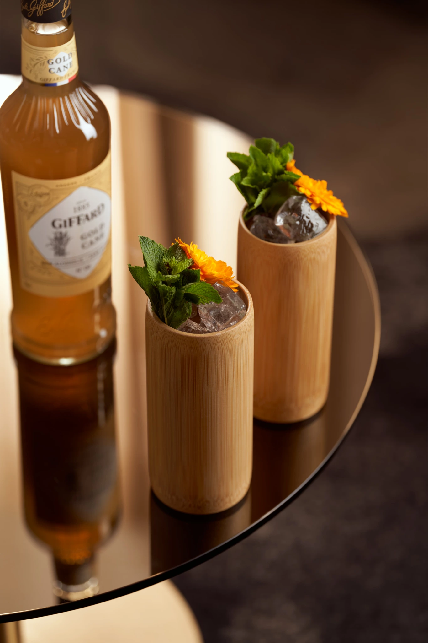 Giffard Gold Cane Bouteille de liqueur Giffard avec deux tasses en bambou contenant des cocktails, de la glace, de la menthe et des fleurs d'oranger sur une table réfléchissante - photographie de nature morte.