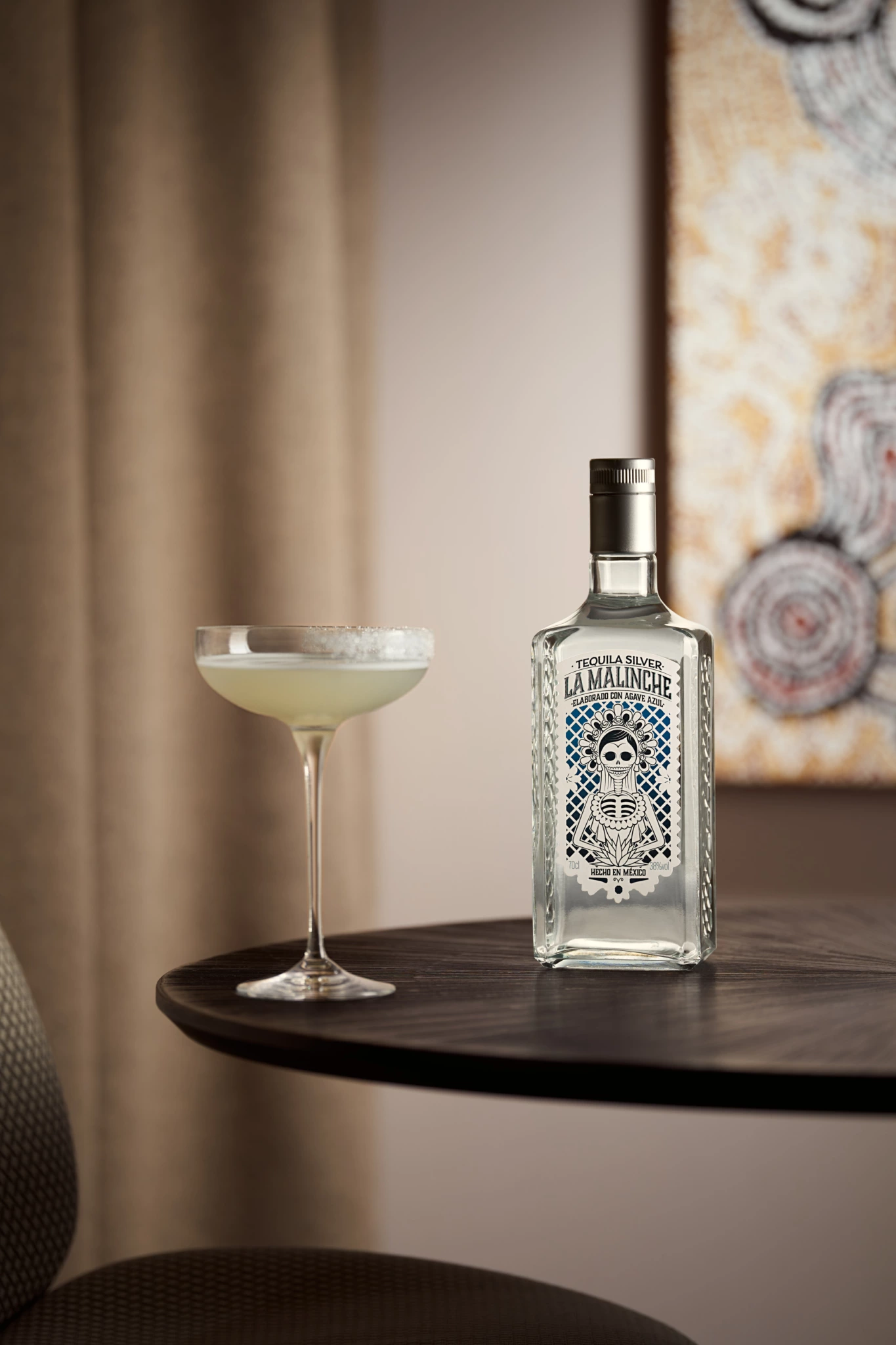 La Malinche Bouteille de tequila La Malinche et verre à coupe avec un cocktail pâle sur une table sombre ; peinture floue et rideau beige en arrière-plan.