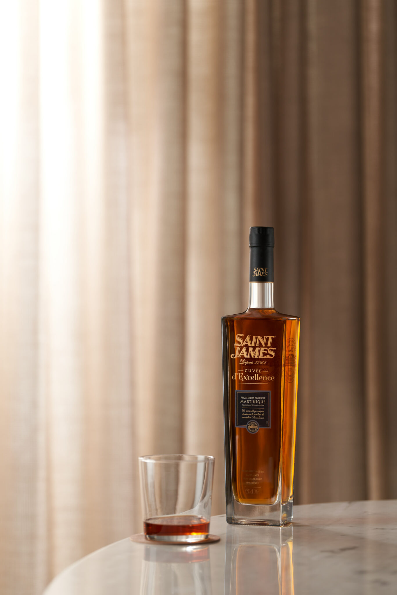 Saint James Excellence Bouteille et verre de rhum Saint James avec un liquide ambré sur une surface en marbre, photographiés avec un éclairage de boîte à lumière et des rideaux neutres derrière.