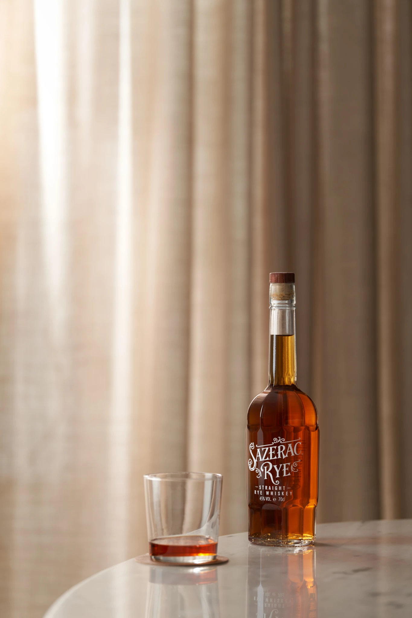 Sazerac Rye Bouteille et verre de whisky Sazerac Rye sur marbre, placés contre des rideaux beiges uniformément éclairés, dans une configuration contrôlée de nature morte en studio.