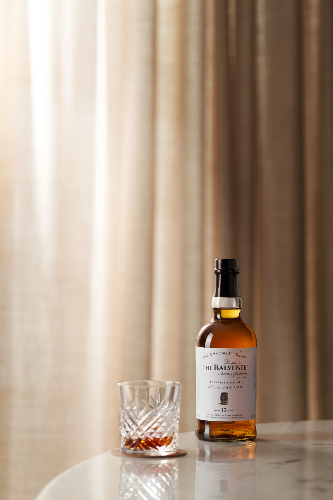 The Balvenie La bouteille Balvenie DoubleWood 12 et un verre en cristal rempli sur du marbre, avec des rideaux beiges derrière, photographiés comme une nature morte de studio.