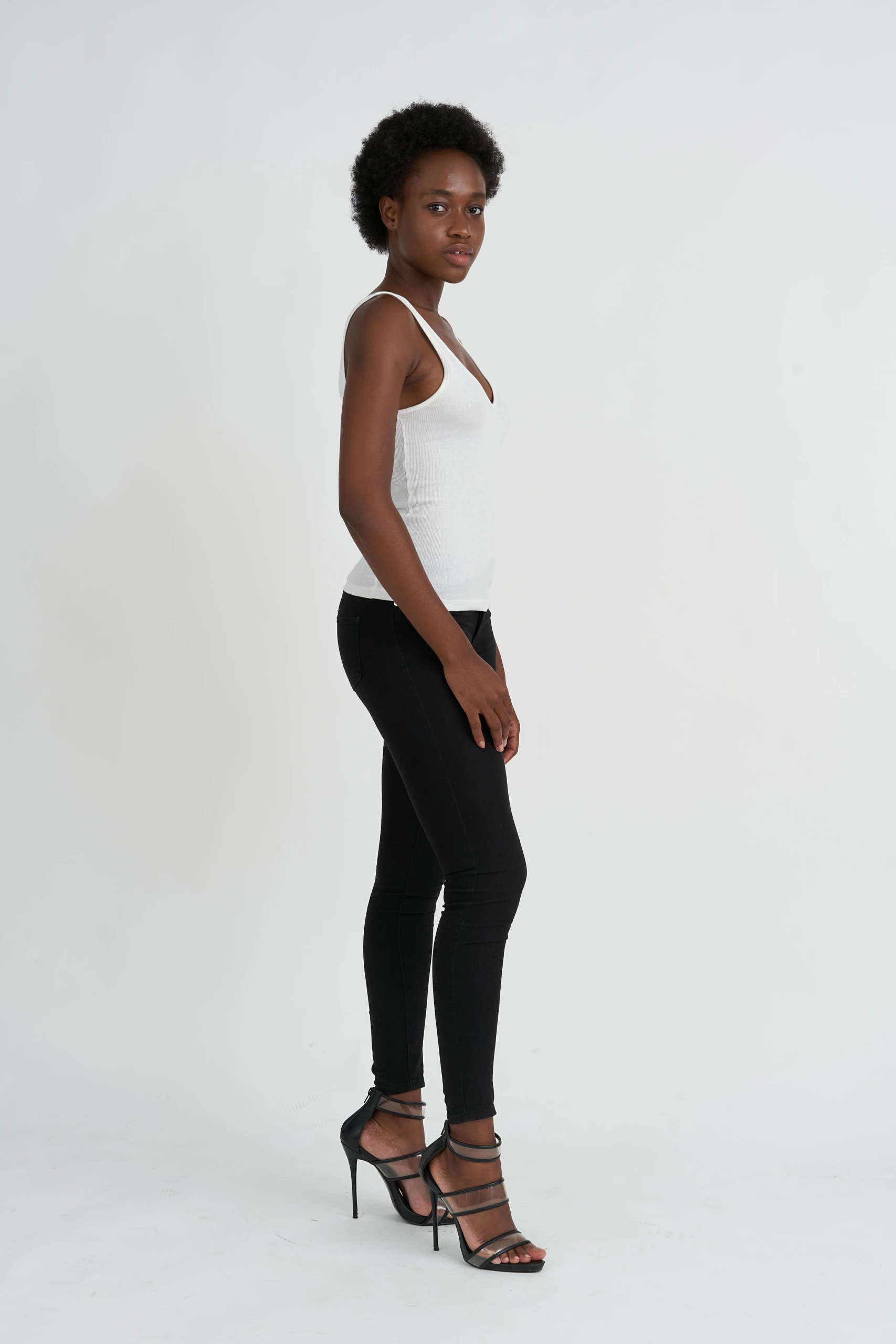Une femme se tient de profil sur un fond blanc sans couture, vêtue d'un débardeur blanc, d'un legging noir et de talons pour un portrait en studio.