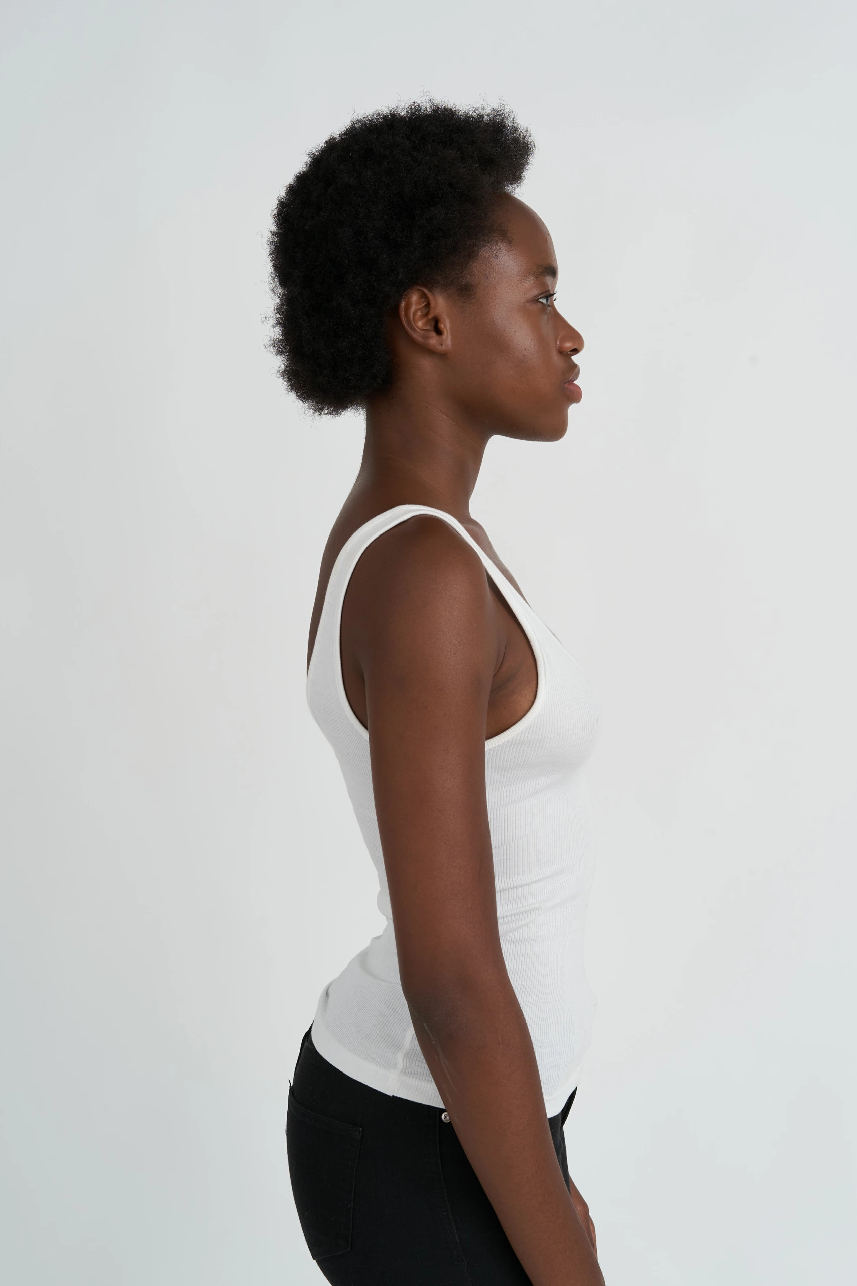Une personne de profil, tournée vers la droite, portant un haut blanc sans manches et un pantalon noir, sur un fond de studio blanc sans couture.